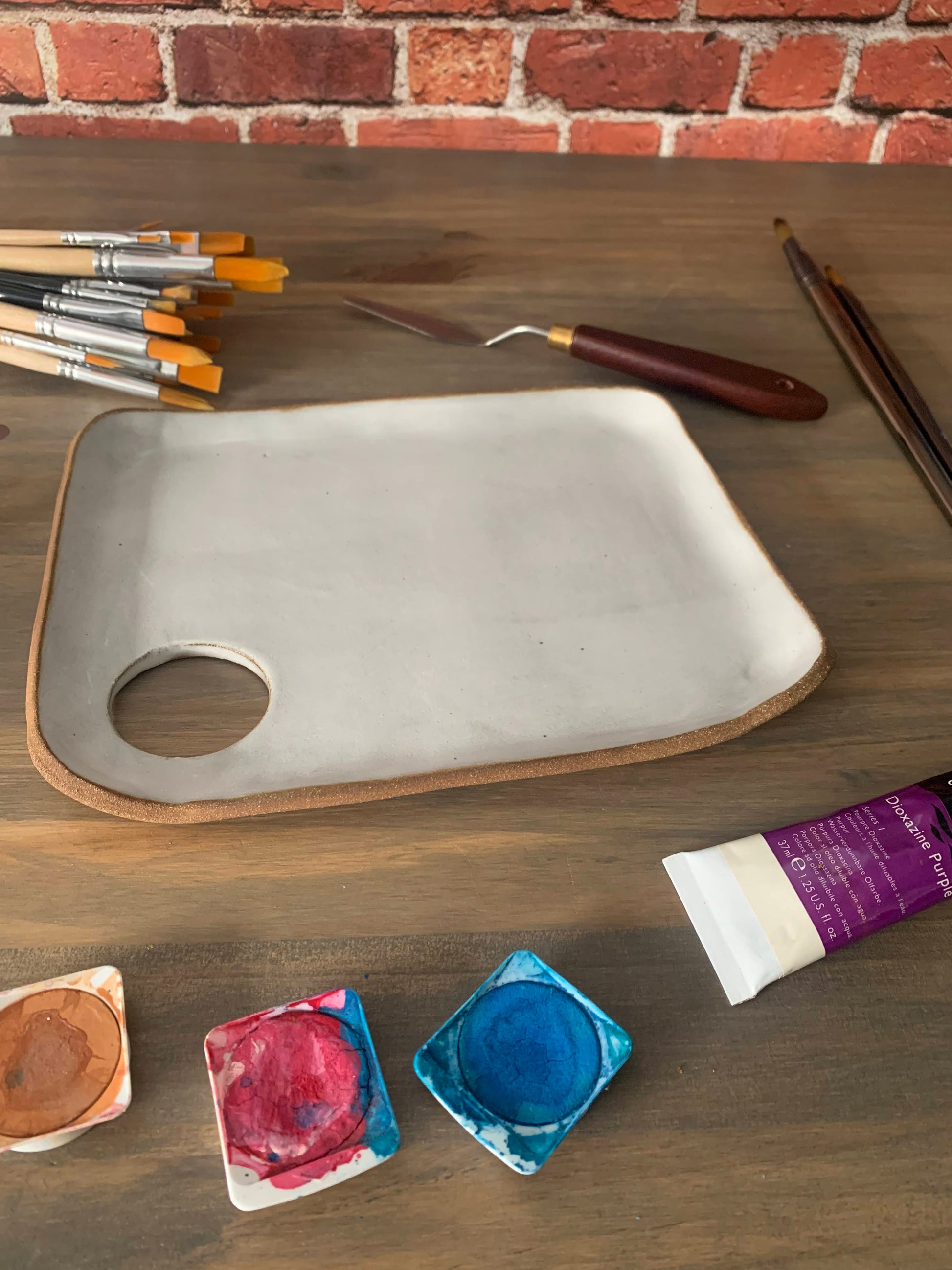 Art supplies including brushes, a handmade ceramic palette, and paint tubes on a wooden surface with a brick wall background.