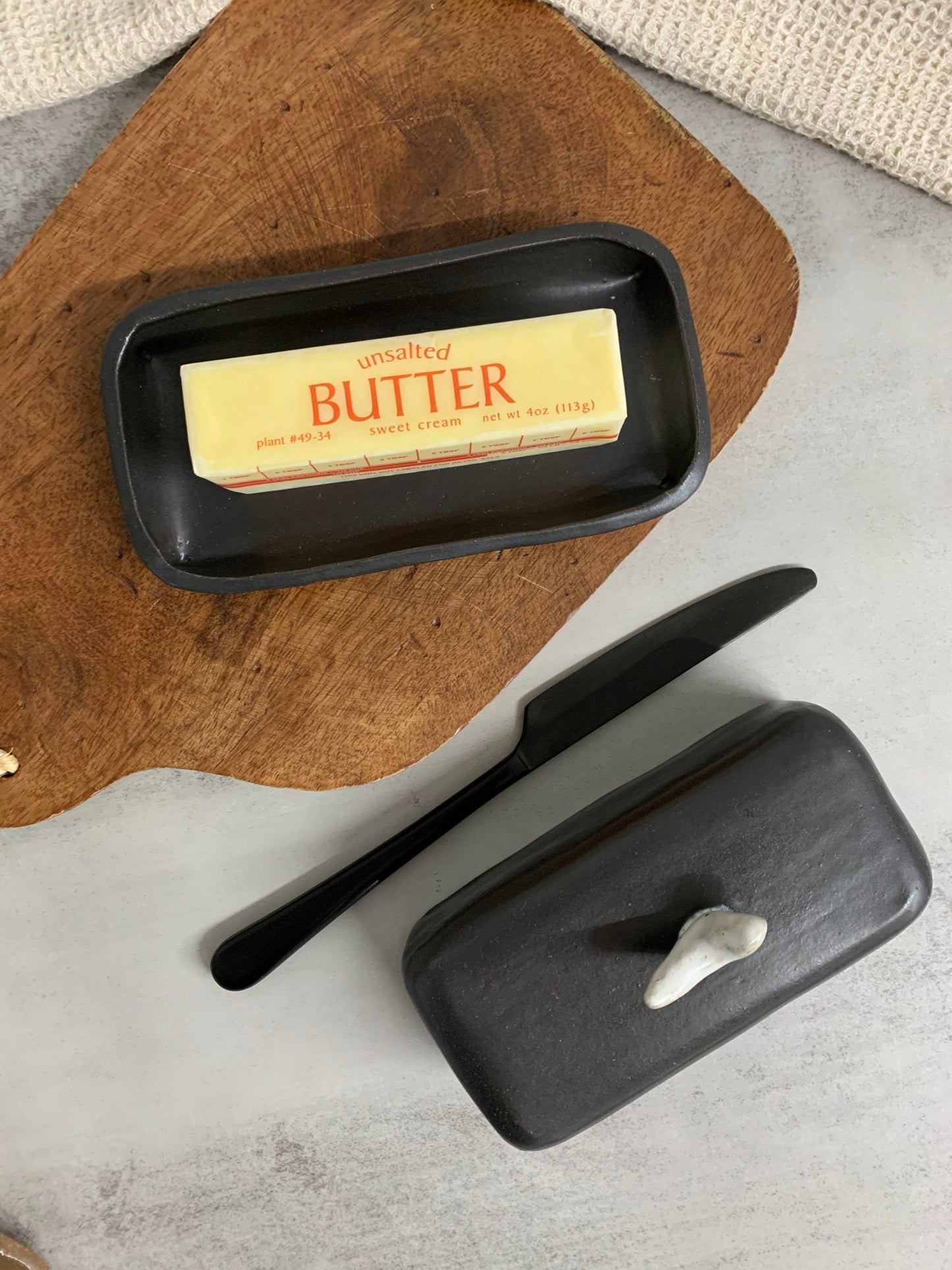 Black handmade ceramic butter dish with a stick of butter, knife, and black lid on wooden board.