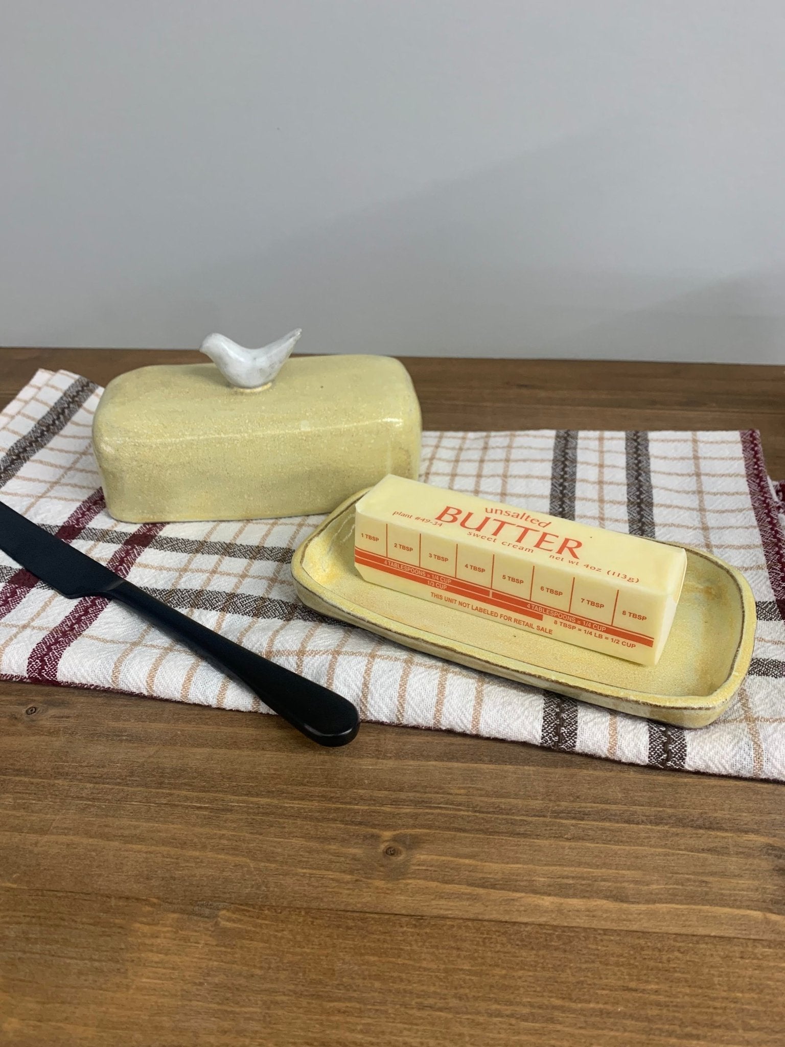 Handmade ceramic butter dish with a bird on top and a stick of butter on a checkered cloth on a wooden surface.