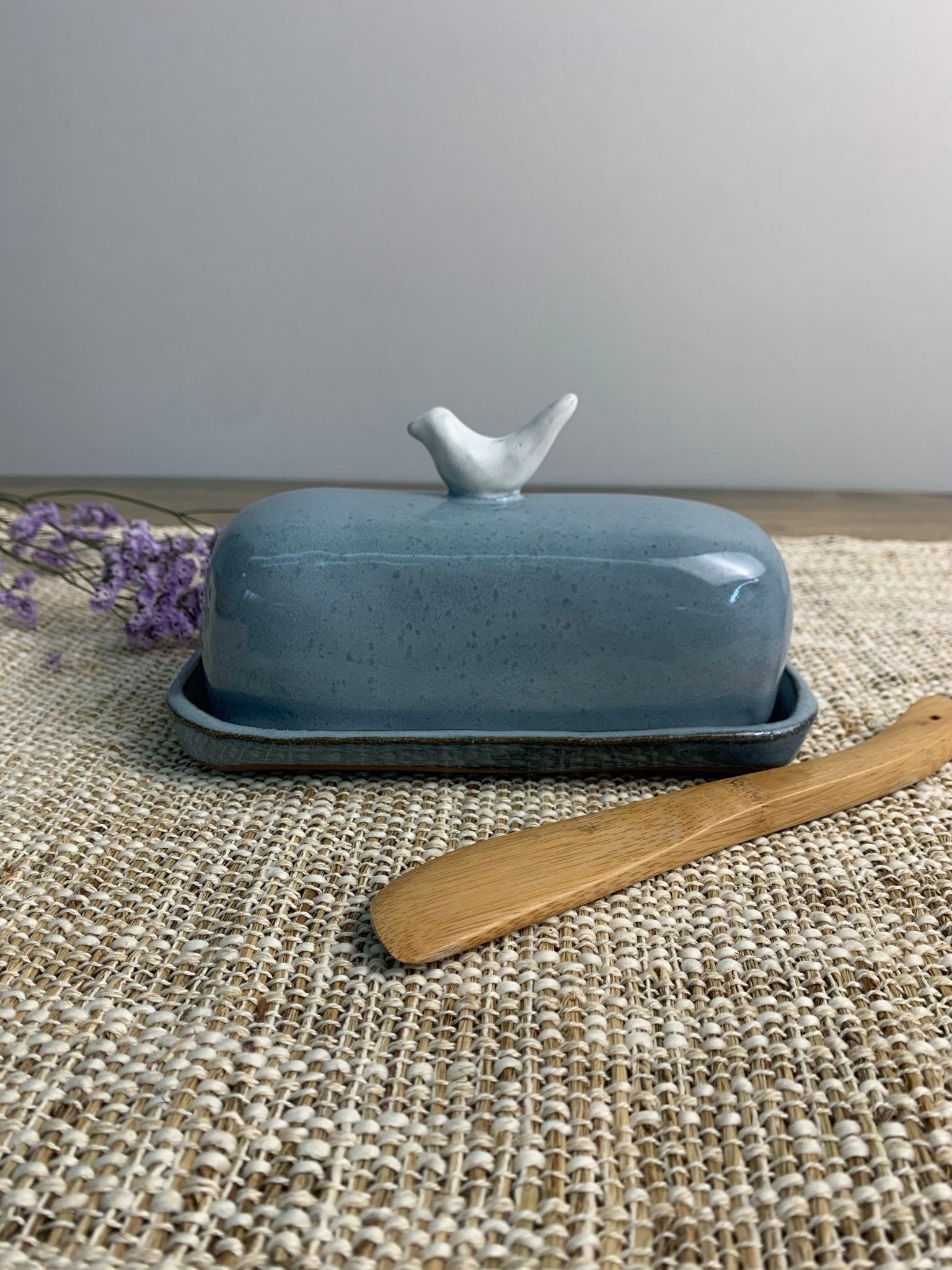 Blue ceramic butter dish with bird design on lid and wooden knife on woven mat