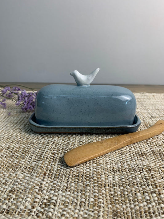 Blue ceramic butter dish with bird design on lid and wooden knife on woven mat