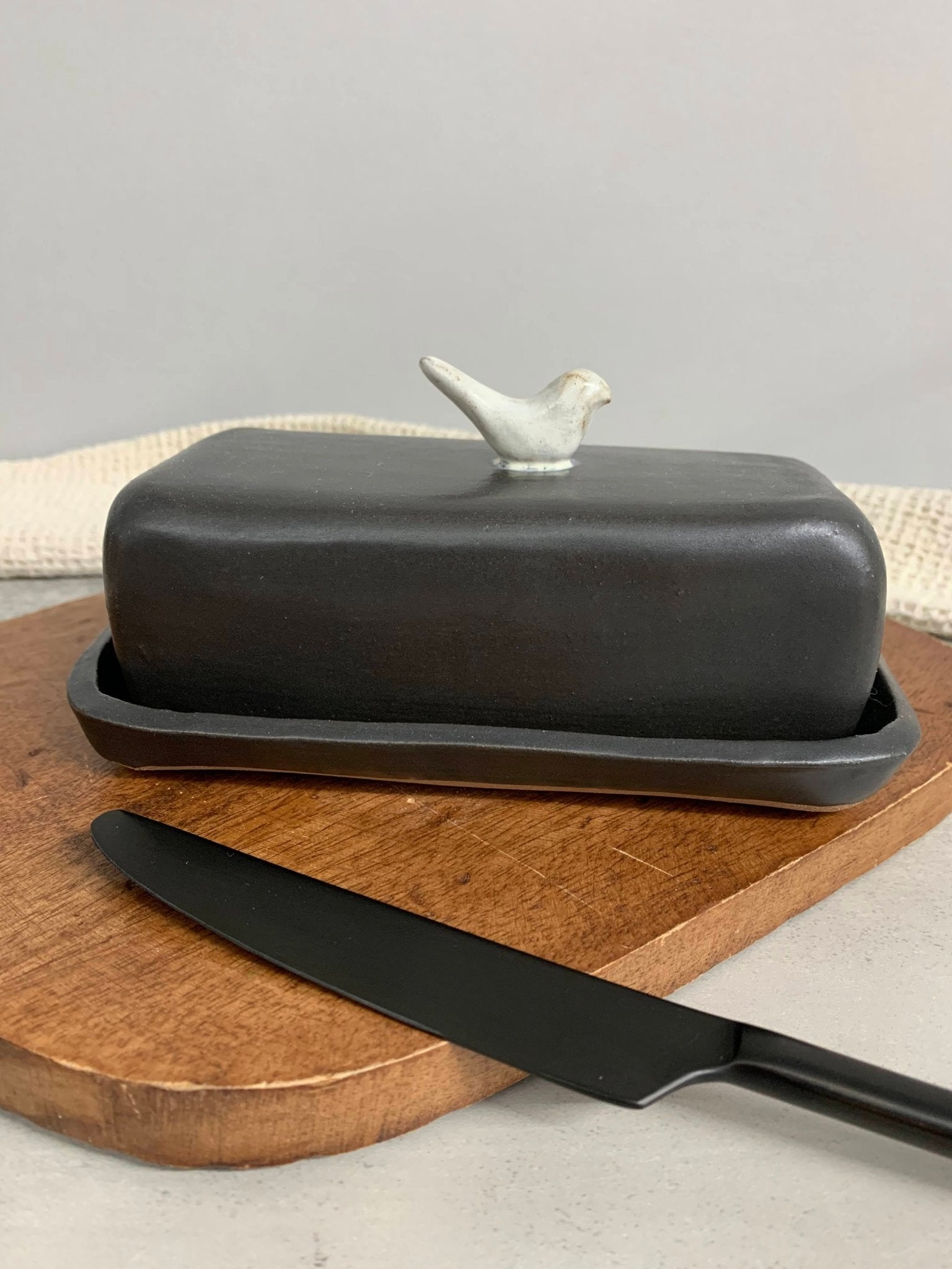 Black handmade ceramic butter dish with bird design on lid and black knife on wooden board