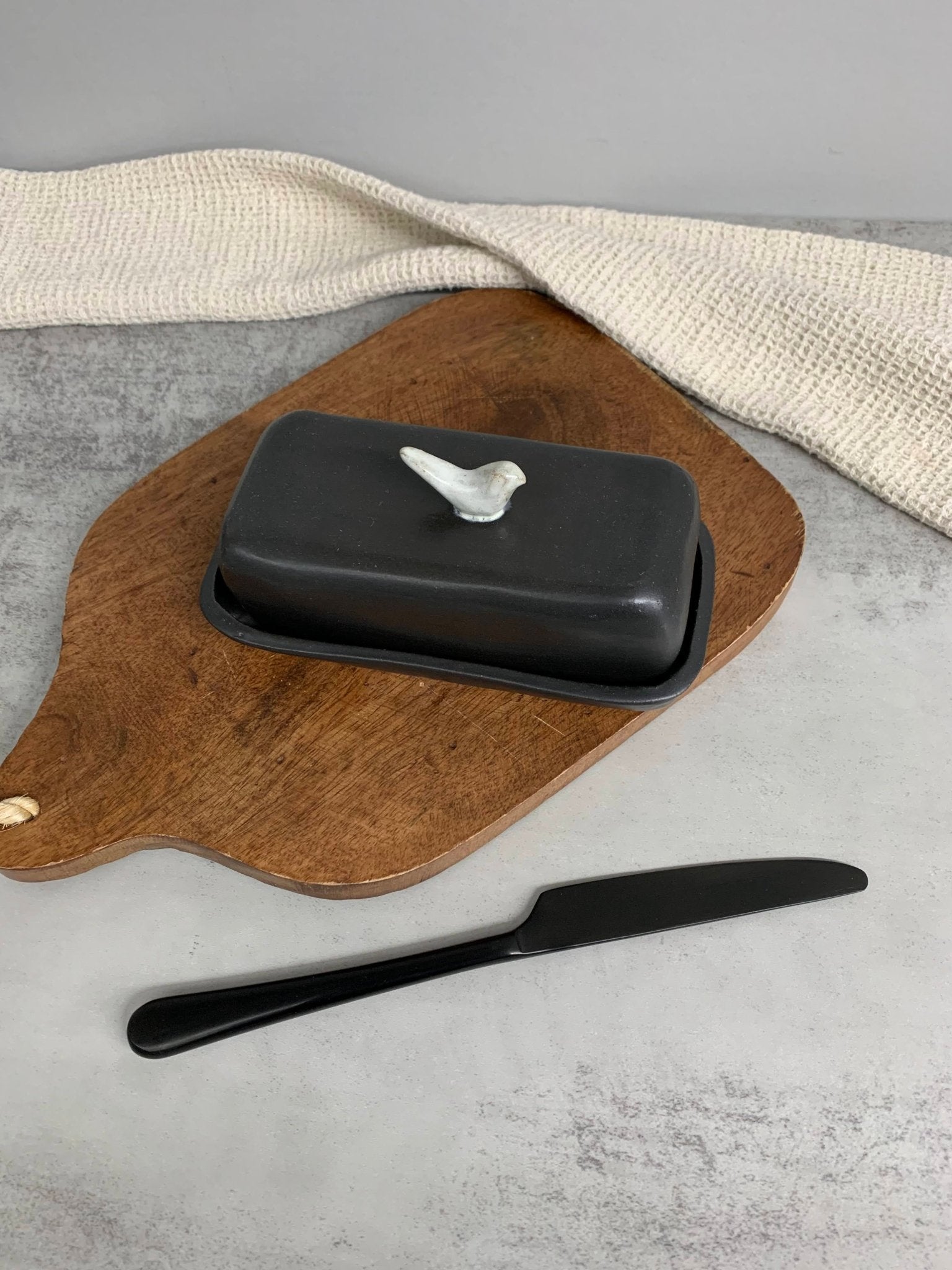 Black handmade ceramic butter dish with bird design on a wooden board with a knife on a gray surface.