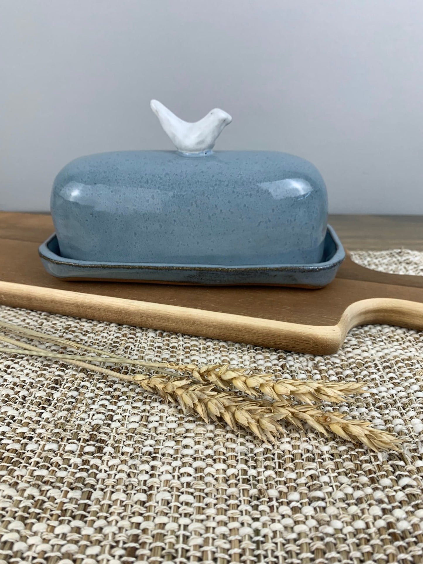 Blue ceramic butter dish with a white bird on top, wooden knife, and wheat stalks on a woven mat.