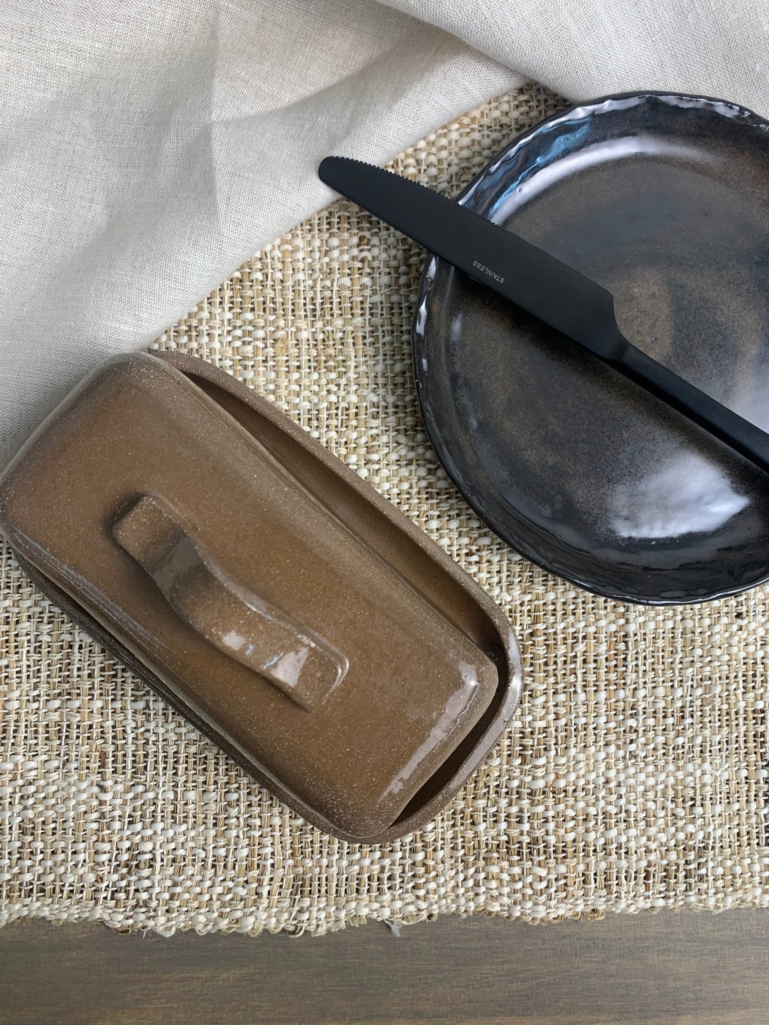 Brown handmade ceramic butter dish with a handle on a textured surface with a black plate and knife.