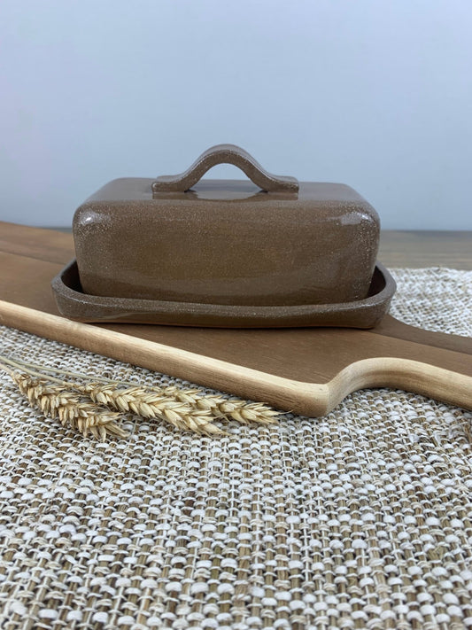 Brown ceramic butter dish with wooden knife and wheat on a textured surface