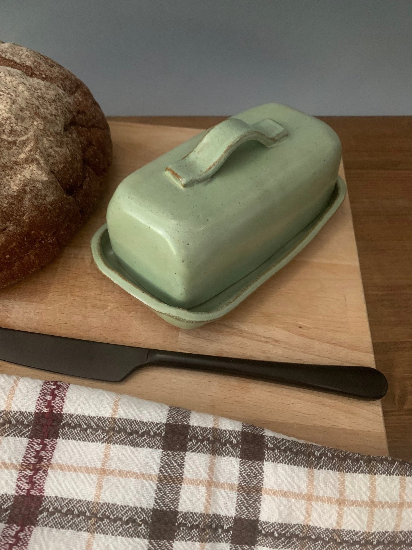 Green handmade ceramic butter dish on a wooden cutting board with bread and a knife, on a checkered tablecloth.