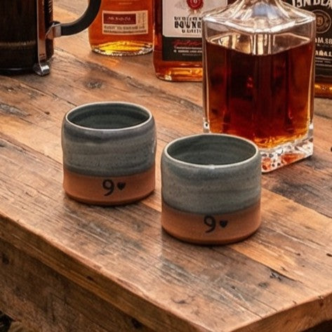 Two ceramic pottery glasses with 9  design on a wooden table with whiskey bottles in the background