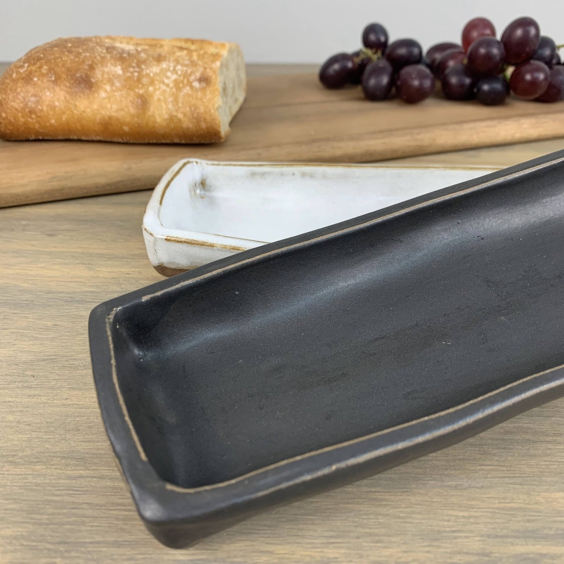 Black handmade ceramic dish on a wooden surface with bread and grapes in the background