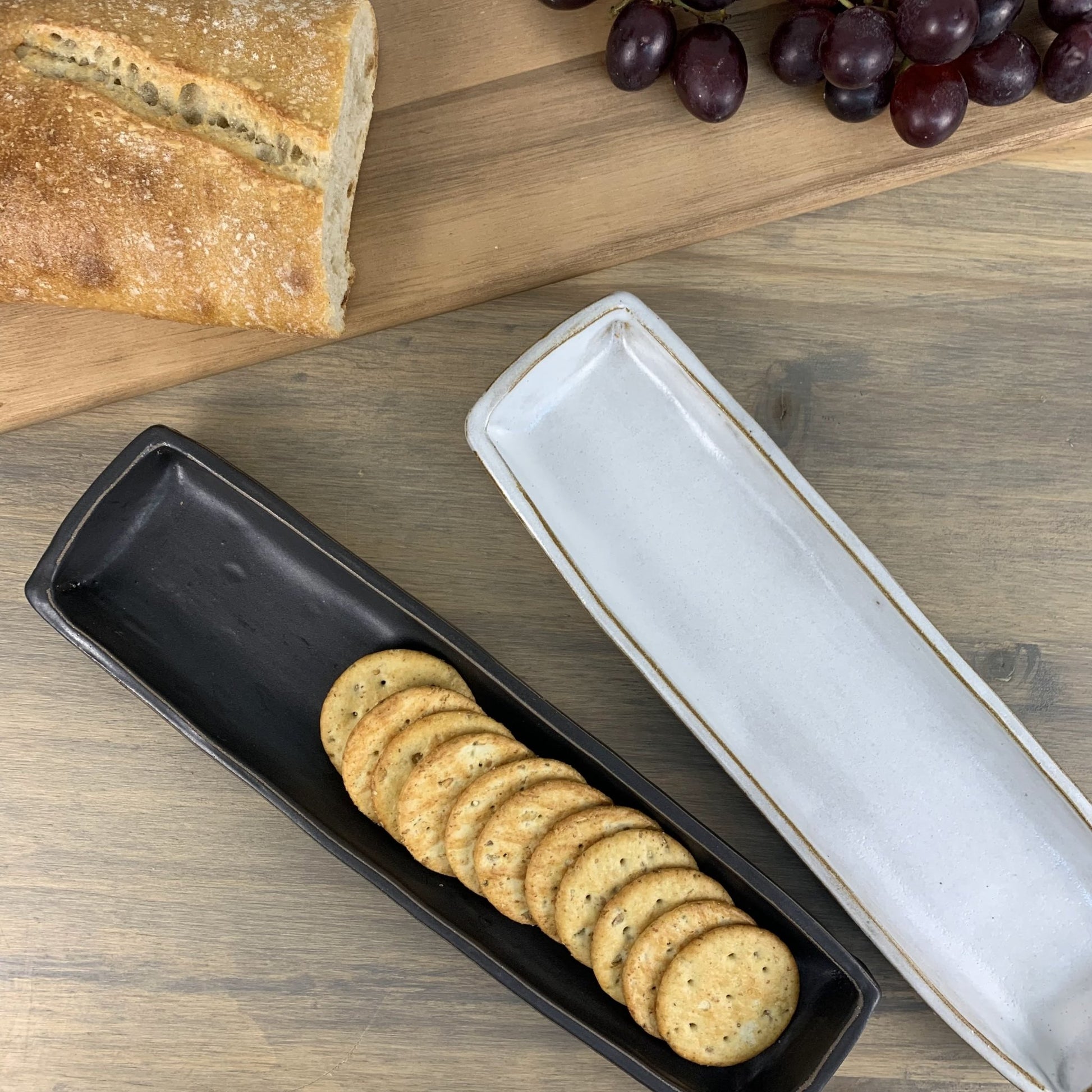 Rectangular black ceramic tray with crackers on a wooden surface with bread and grapes.