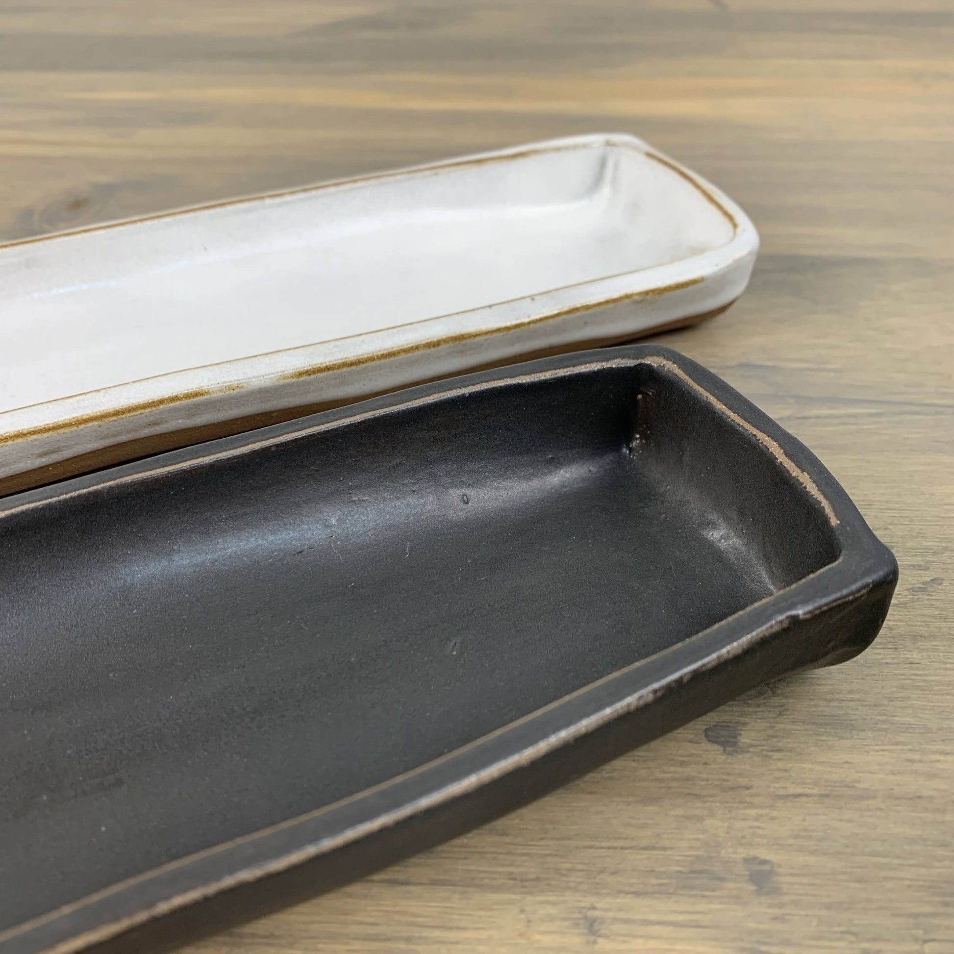 Two ceramic trays, one black and one white with brown edges, on a wooden surface.