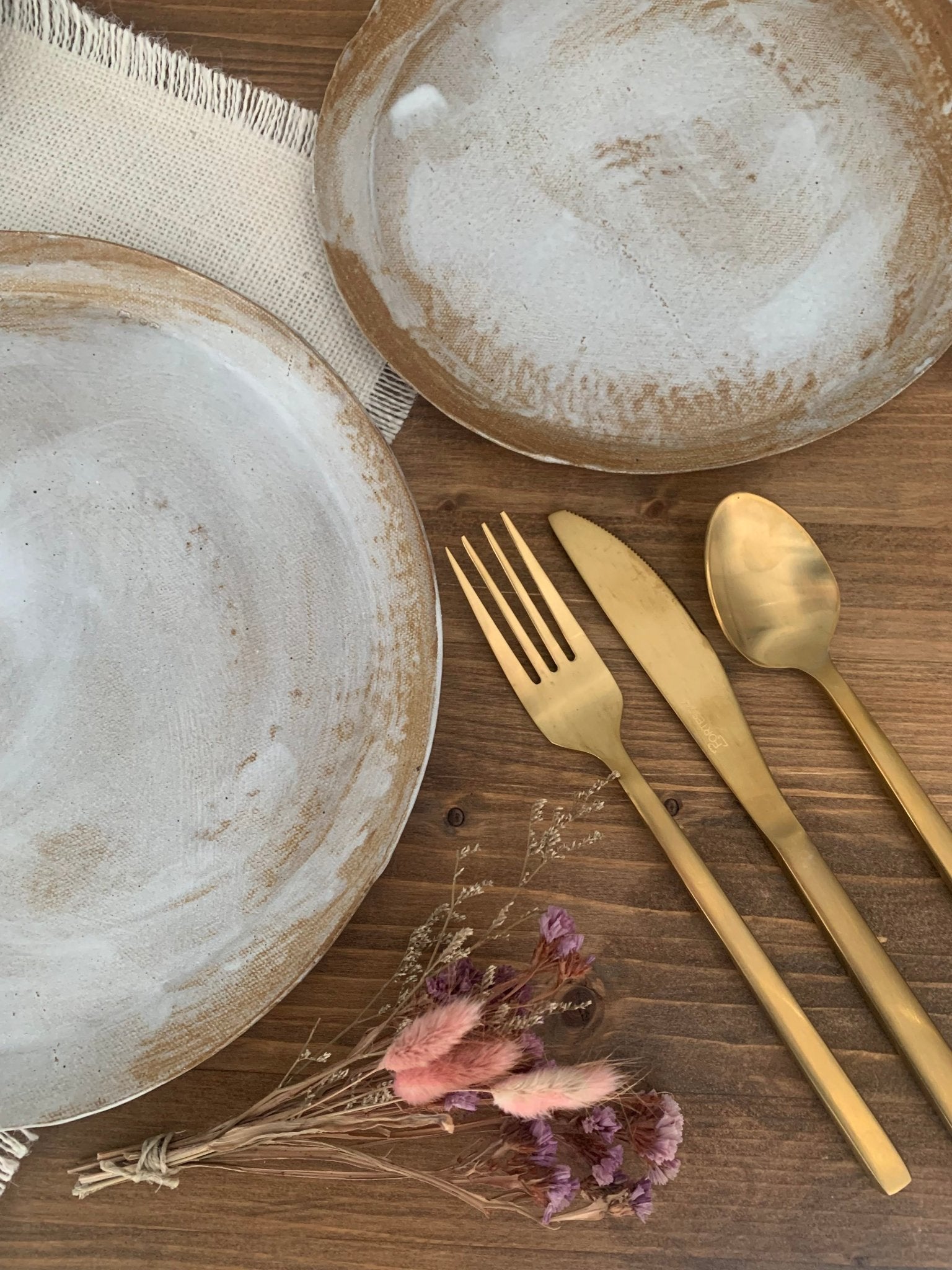 Set of ceramic plates and gold cutlery on a wooden surface with dried flowers.