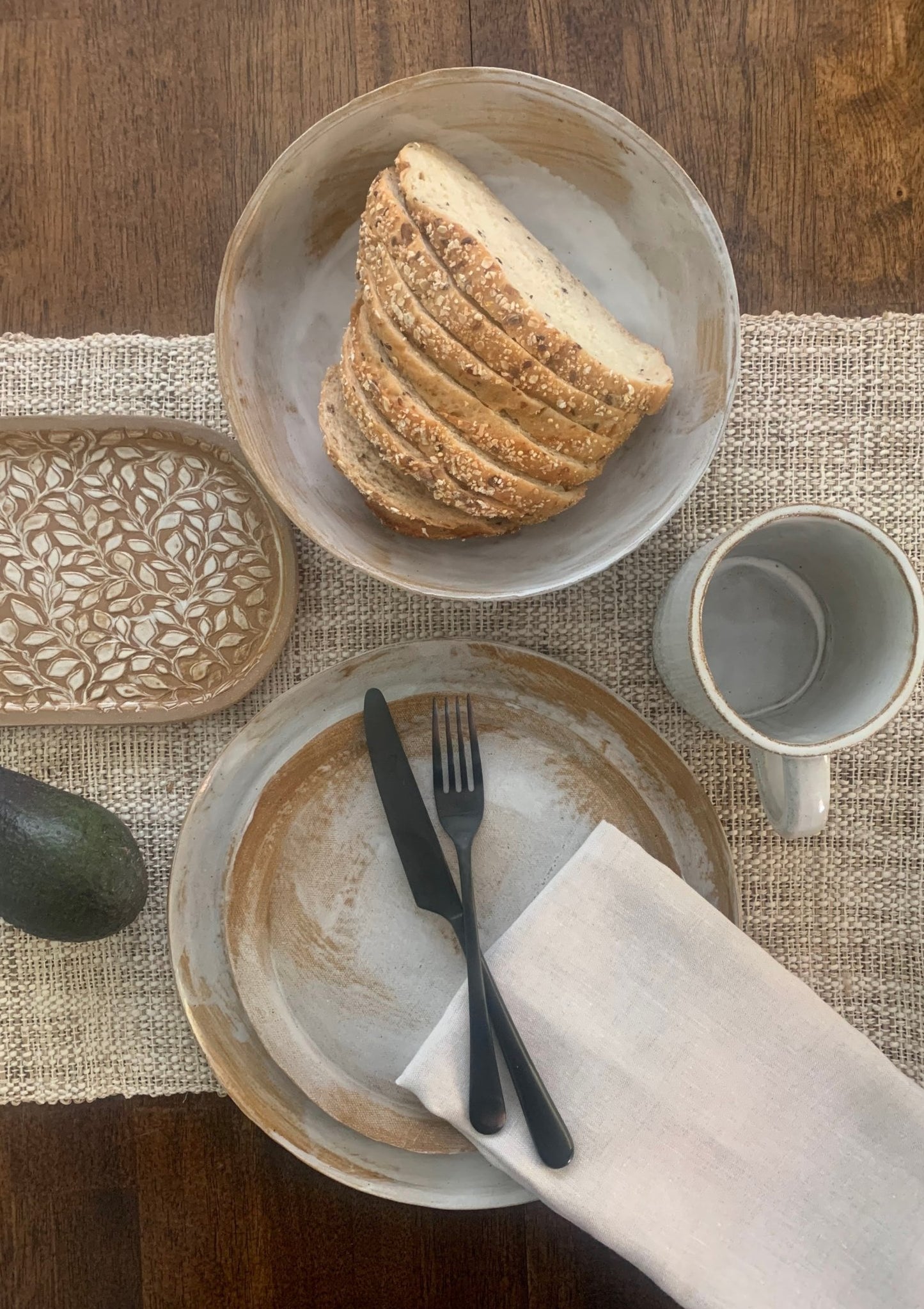Handmade ceramic dinnerware table setting with bread, plates, cutlery, and a mug on a textured tablecloth.
