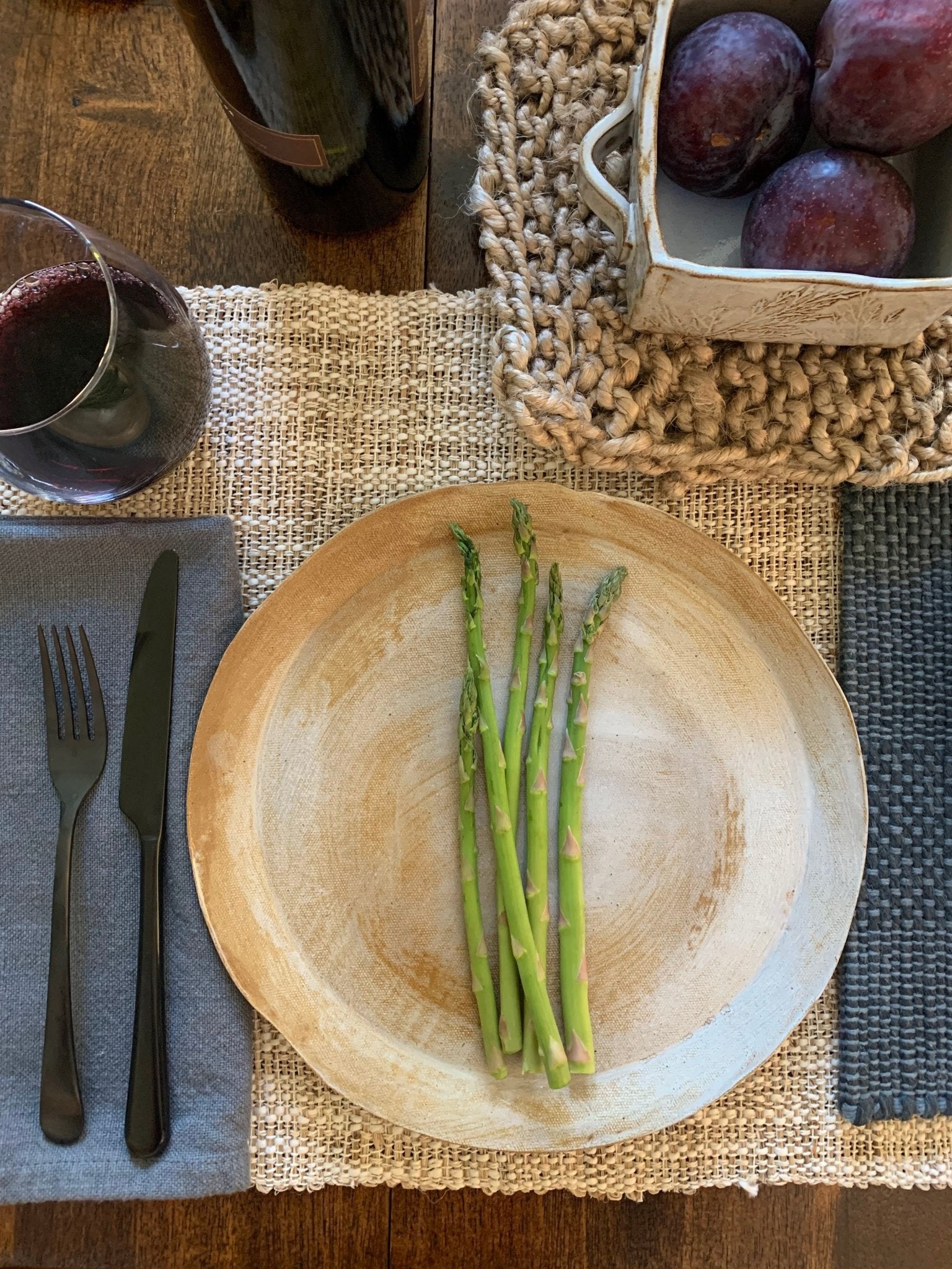 Handmade ceramic plate with asparagus on a table setting with a glass of red wine and a basket of plums.