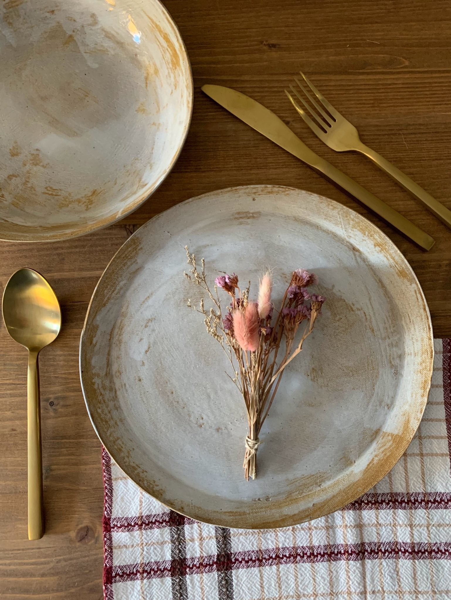 Dinner setting with handmade stoneware, gold cutlery, and dried flowers on a wooden table.
