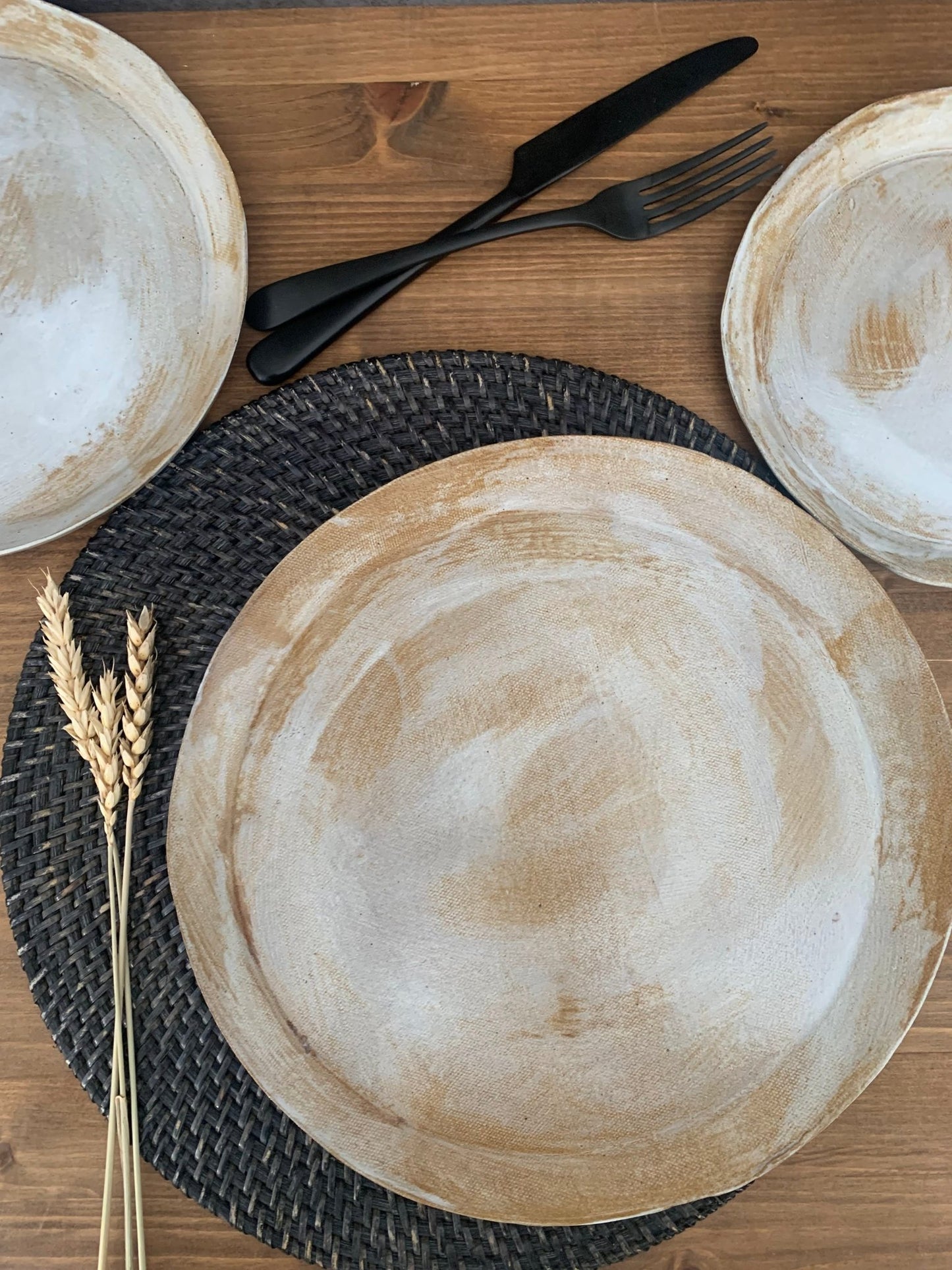 Set of handmade ceramic plates on a woven placemat with black cutlery on a wooden surface.