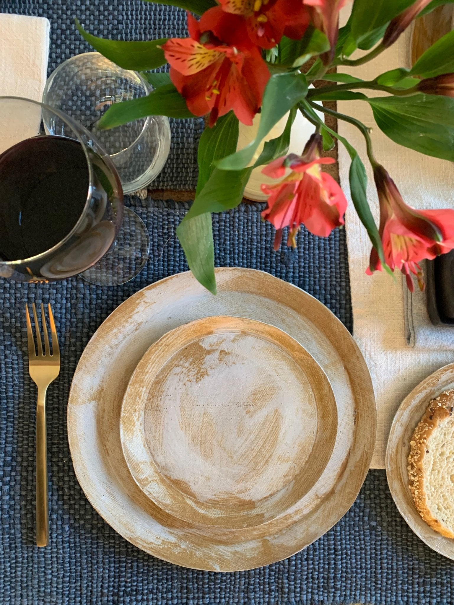 Dining table setting with handmade ceramic dinnerware, cutlery, and a floral arrangement on a blue placemat.