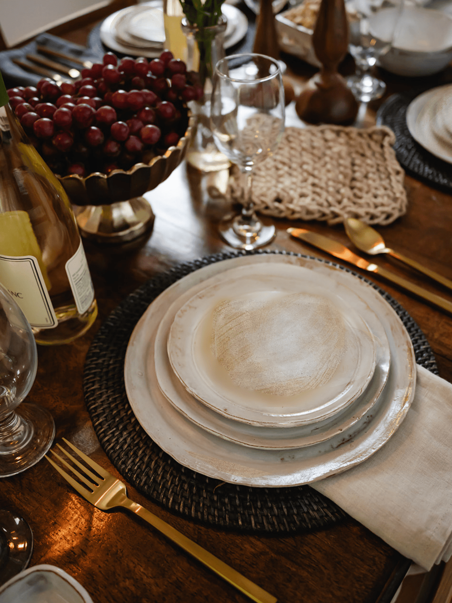 Stack of handmade ceramic stoneware plates with silverware as a place setting on a table with a bowl of grapes, glasses and more dishes