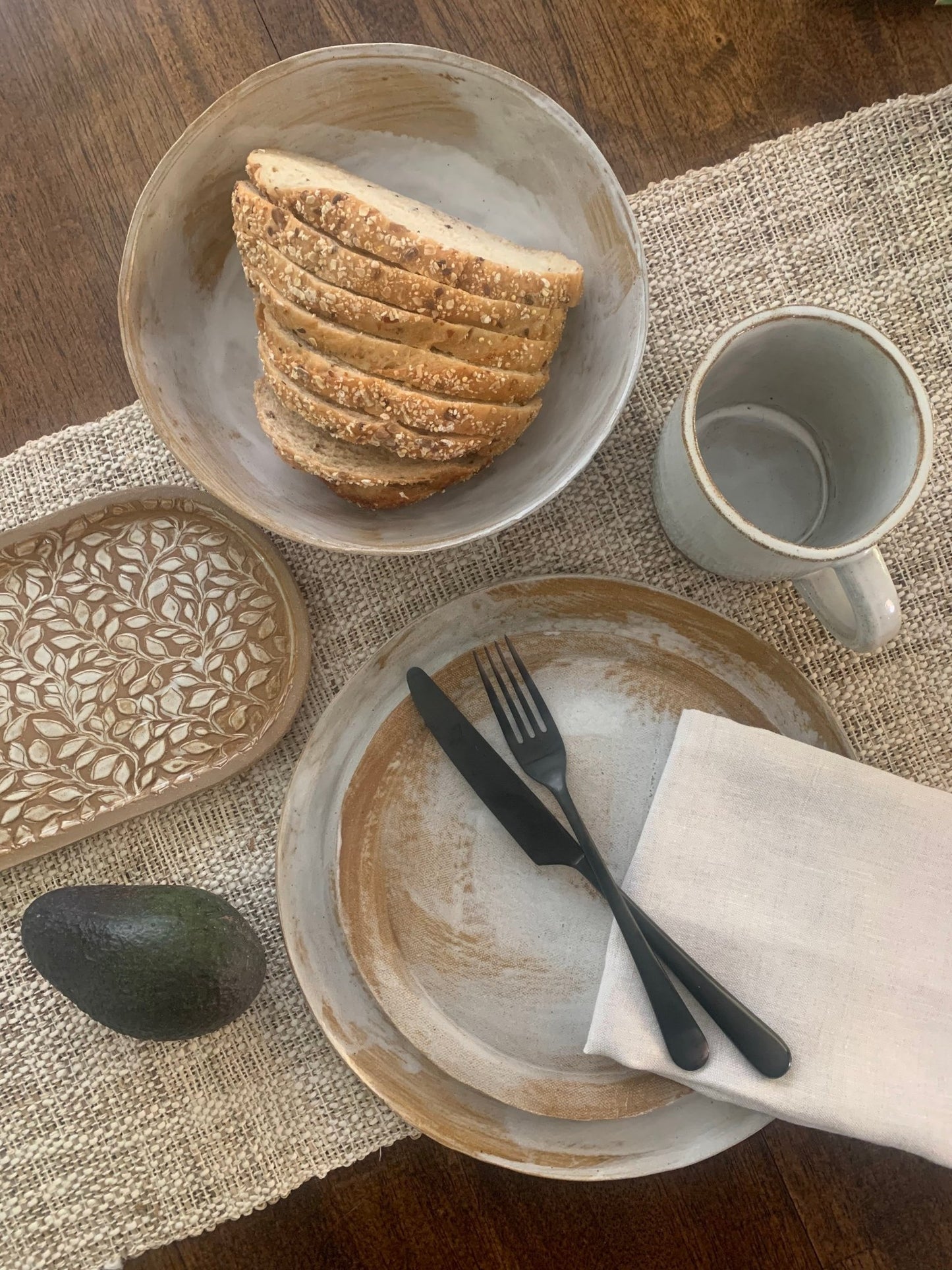 Table setting with handmade ceramic dinnerware, bread, avocado, and cutlery on a textured placemat.