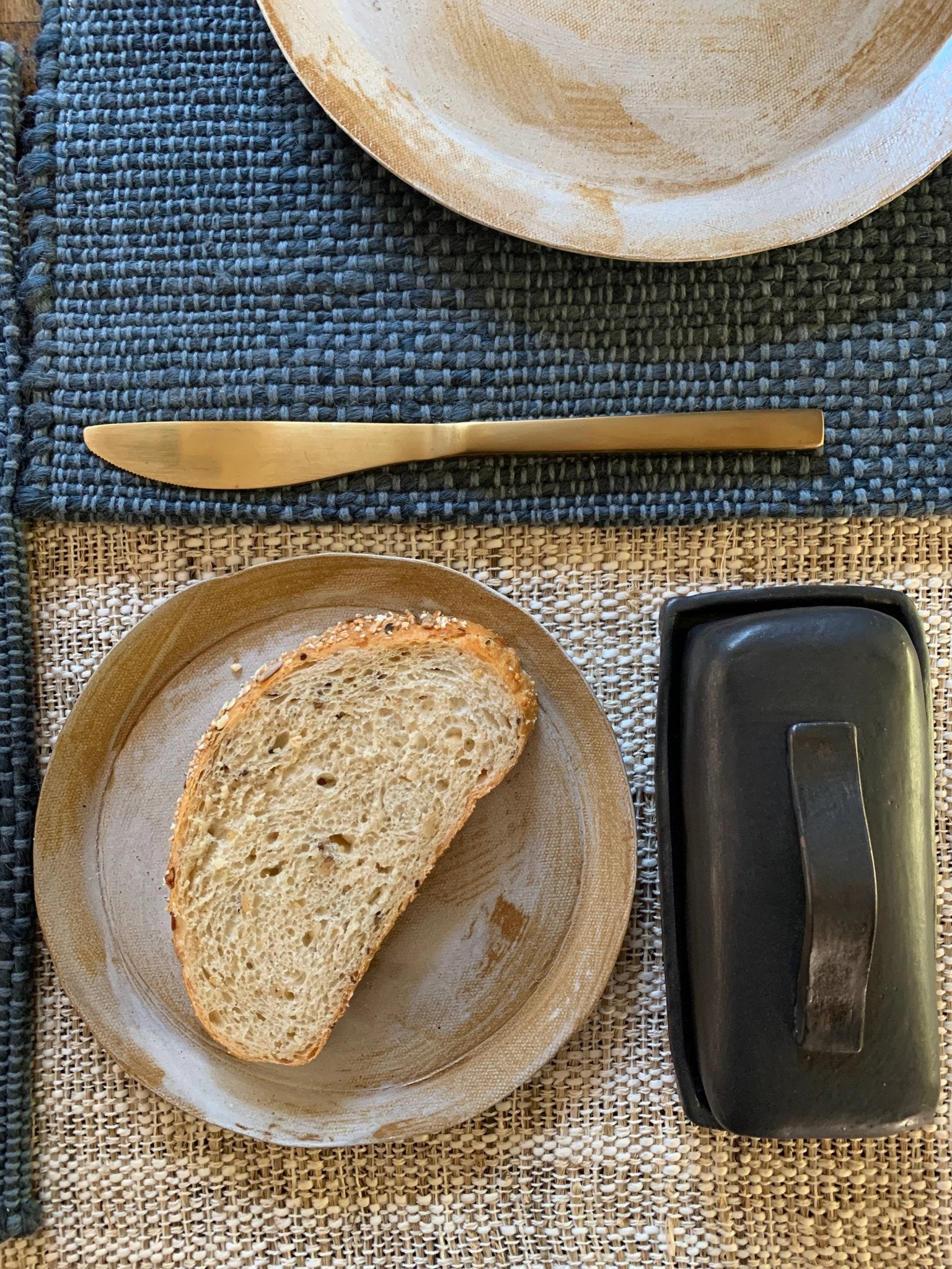 Slice of bread on a handmade plate with a butter dish and knife on a textured surface