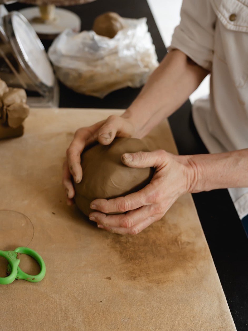 Person shaping a piece of clay on a wooden table with a green tool nearby.