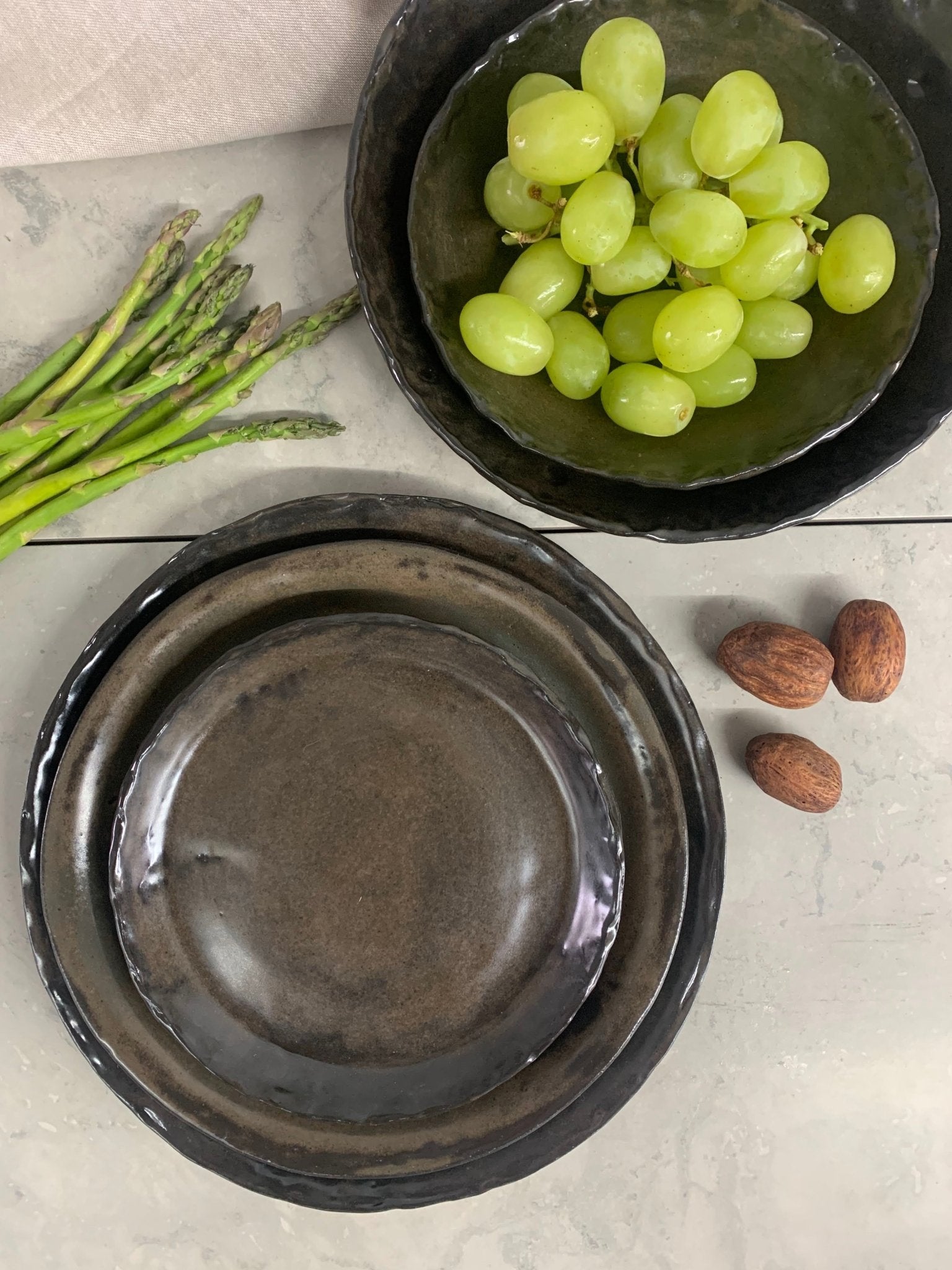 Three dark ceramic plates with a bowl of green grapes and asparagus on a light surface.