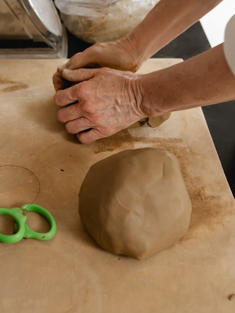 Person kneading clay on a wooden surface with a green tool nearby.