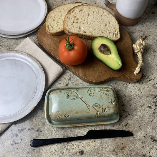 artisan botanical Butter dish with bread, tomato, and avocado on a wooden cutting board.