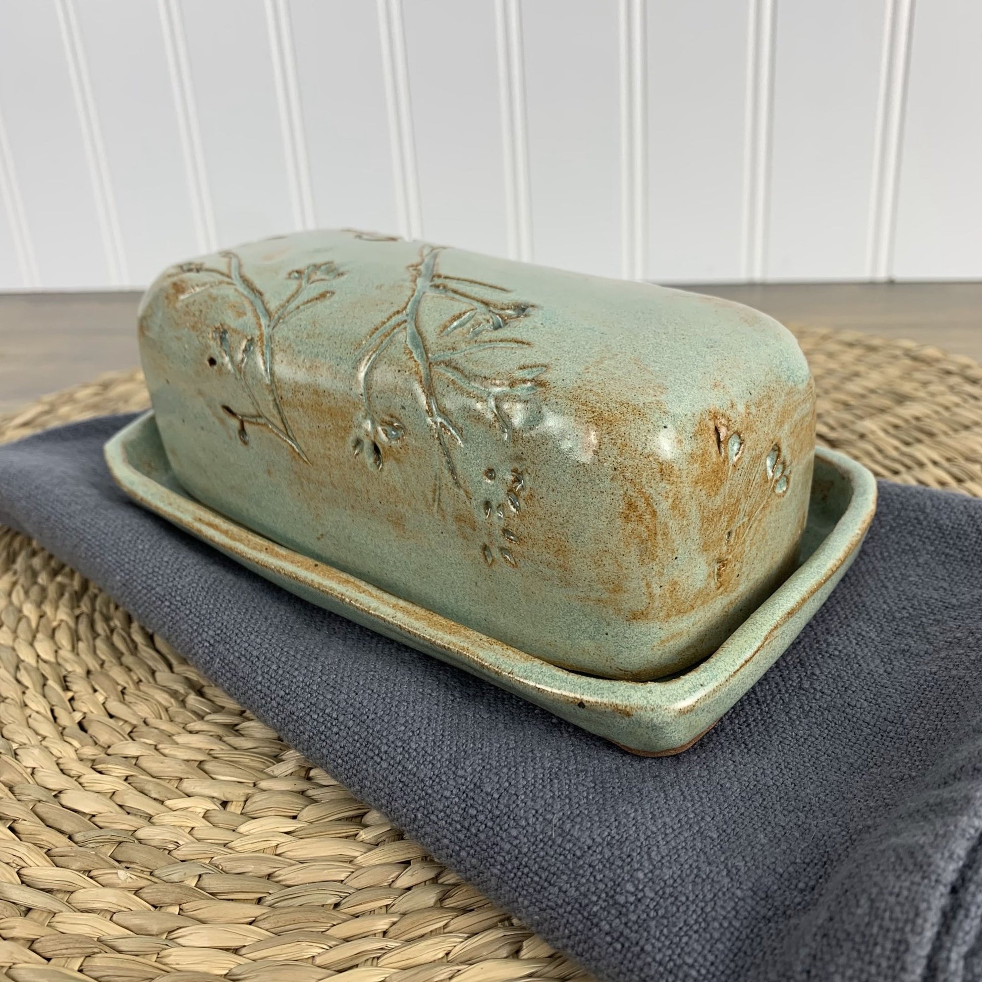 Green handmade ceramic butter dish with floral design on a woven mat and gray cloth