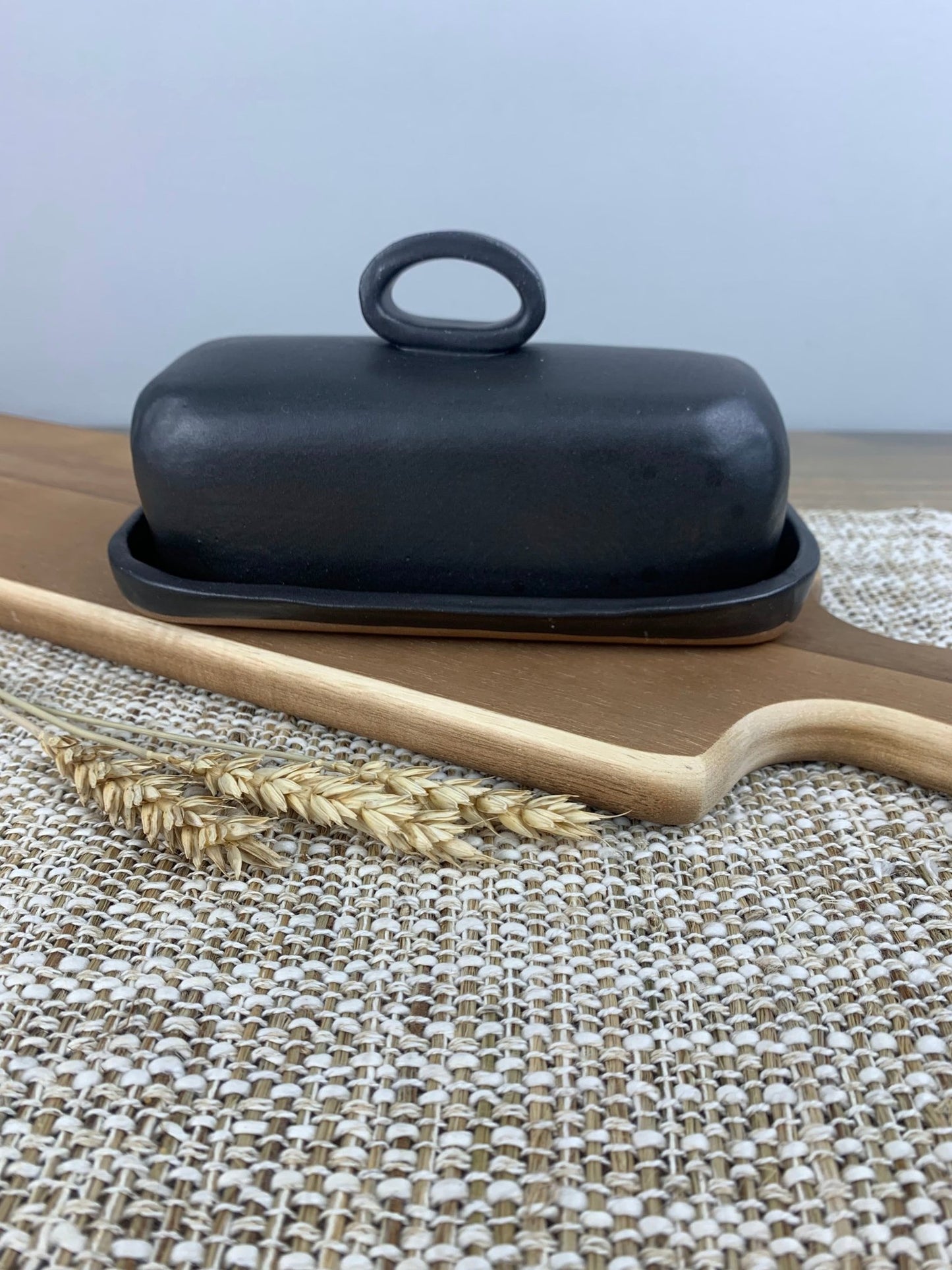 Handmade ceramic black butter dish with a handle on a woven mat with a wooden spoon and wheat stalk.
