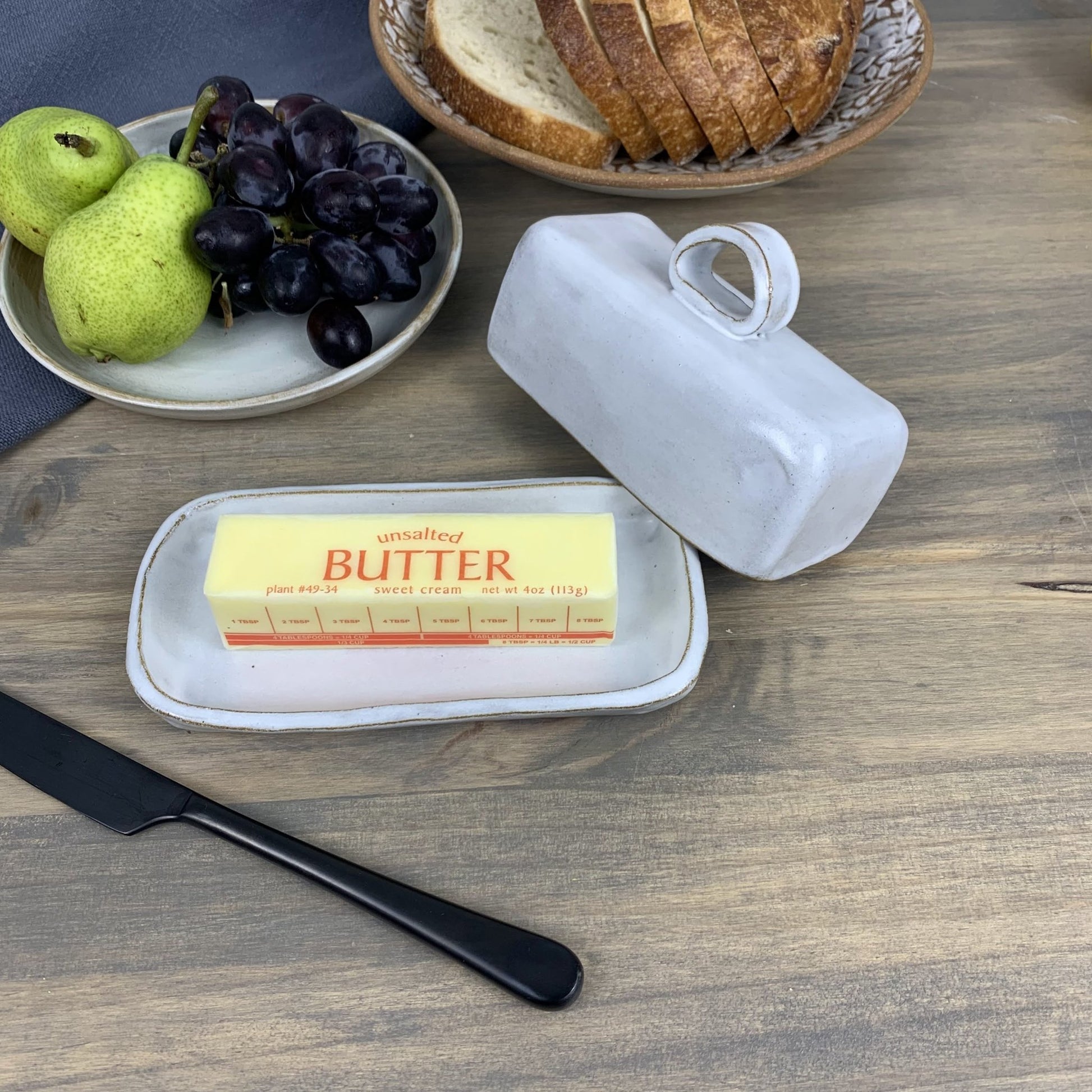 Handmade ceramic butter dish with a stick of butter, knife, grapes, pears, and bread on a wooden surface.