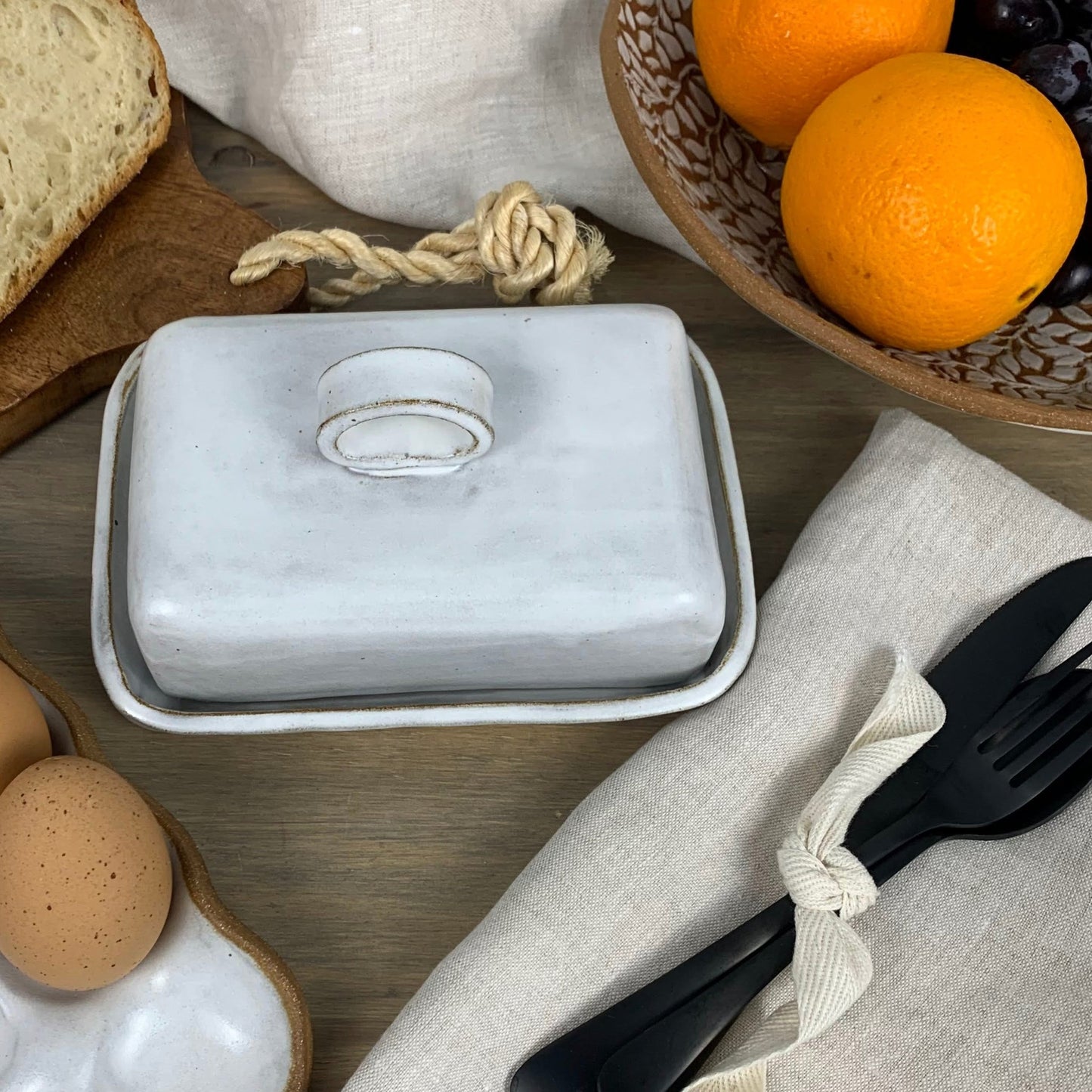 White handmade ceramic butter dish on a wooden surface with bread, oranges, and utensils.