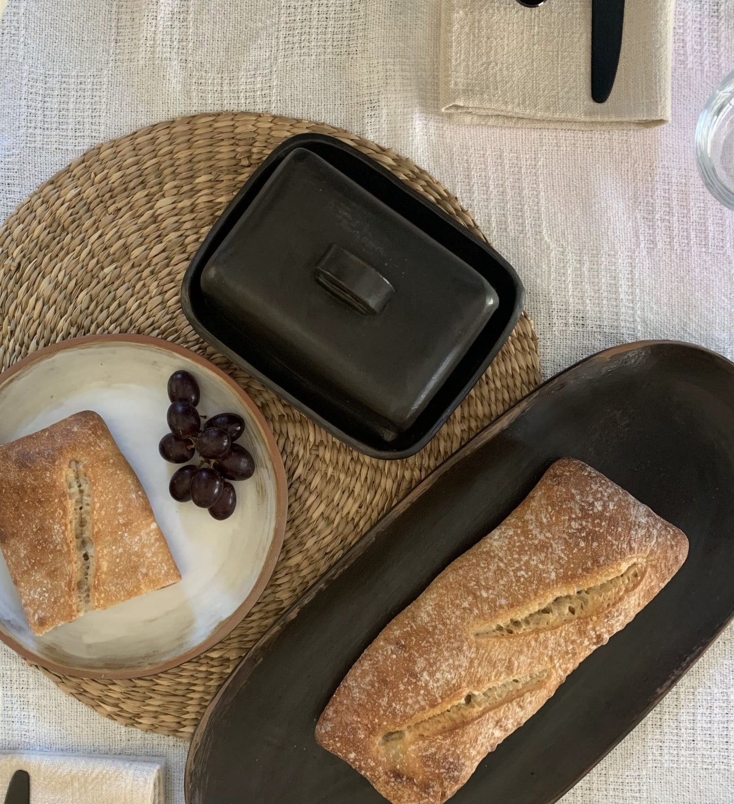 Loaf of bread on a dark handmade ceramic baking dish with a woven mat and plate of grapes in the background.