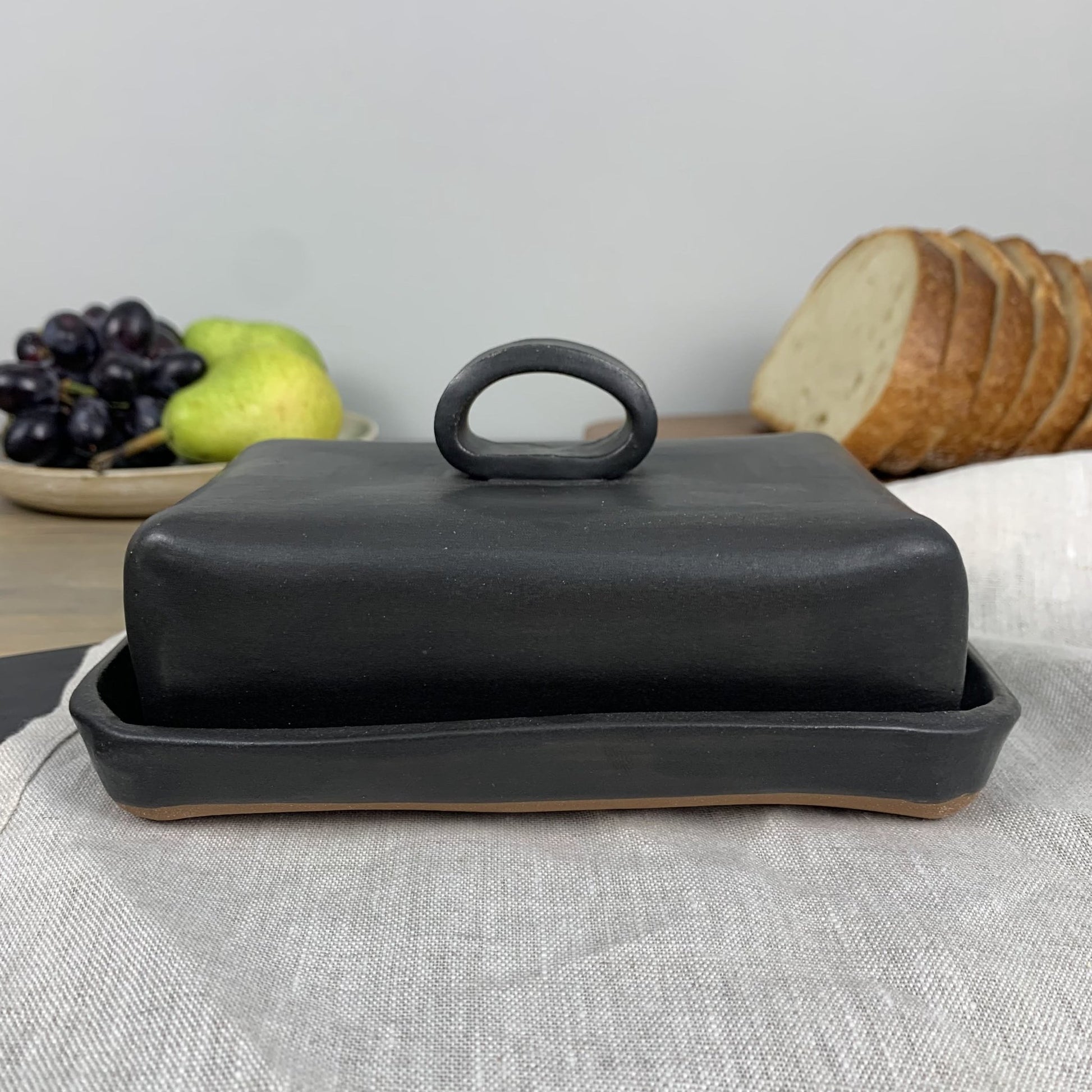 Black handmade ceramic butter dish with lid on a table with fruits and bread in the background