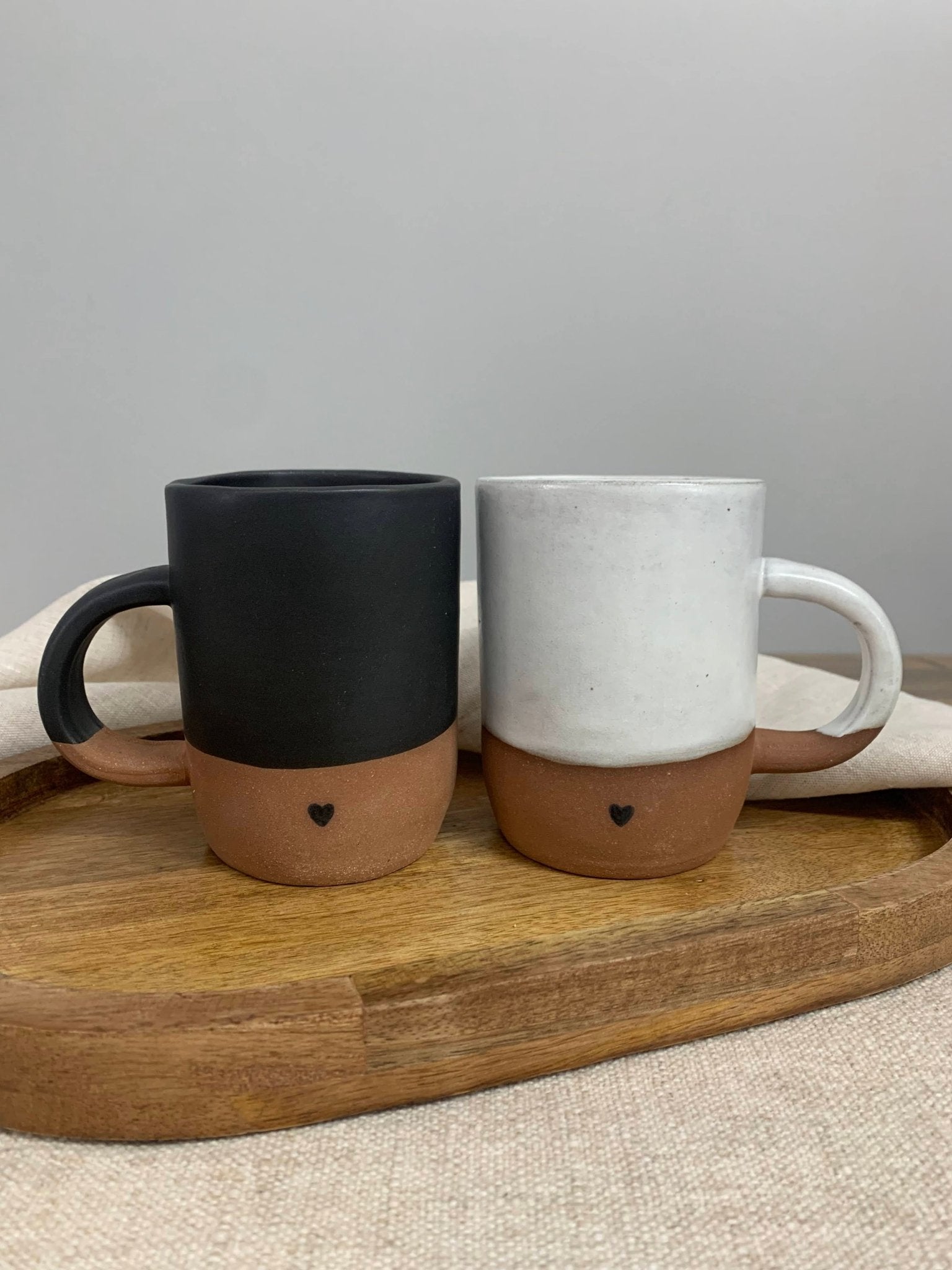 a set of handmade pottery mugs one black and one white with heart images on brown clay bottom sitting on a wood tray with cloth napkin