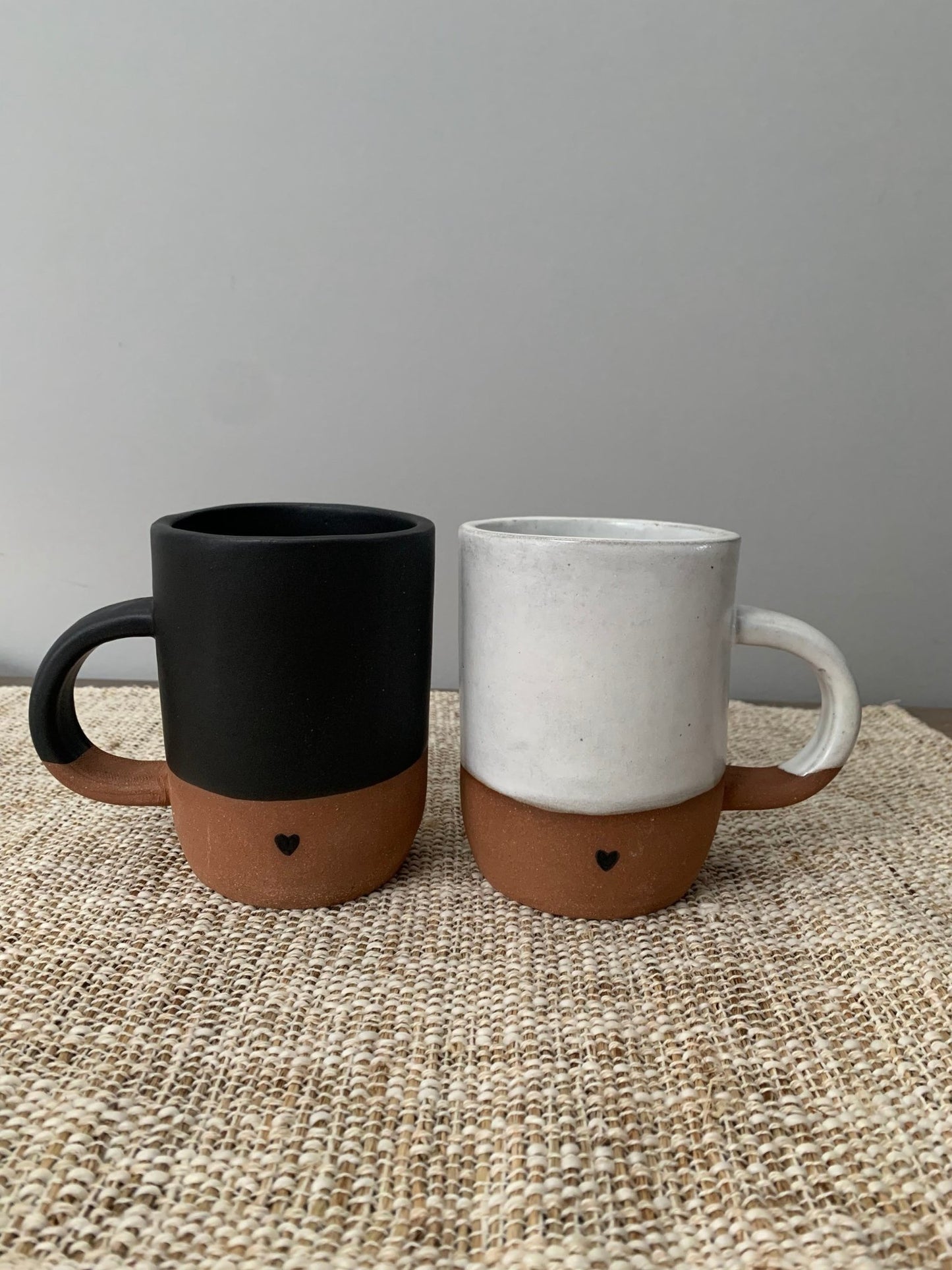 Two artisan ceramic mugs, one black and brown, the other white and brown, on a textured surface with a gray background.
