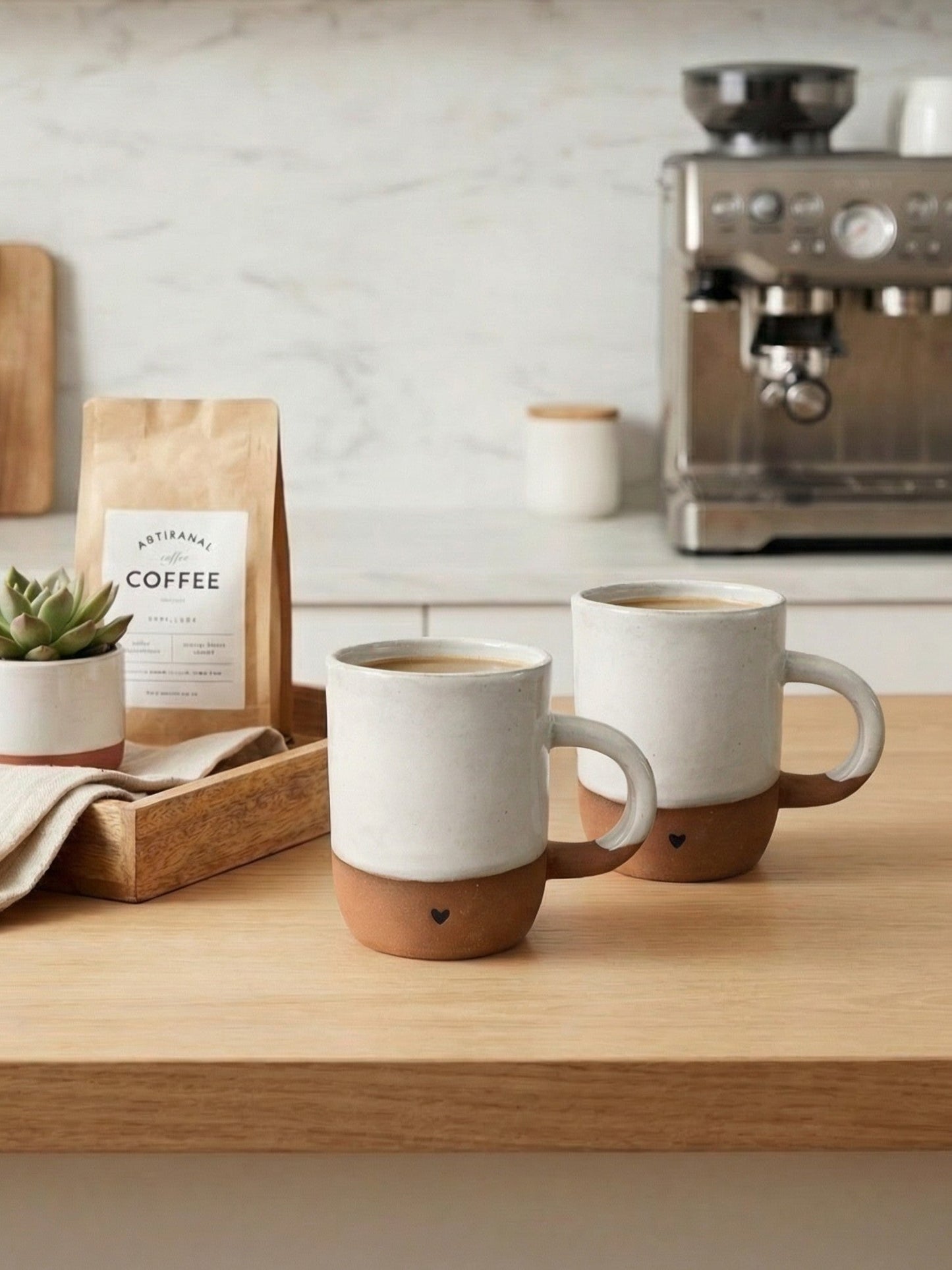 Two ceramic mugs with brown bases with heart design on a kitchen counter with coffee and a coffee machine in the background.