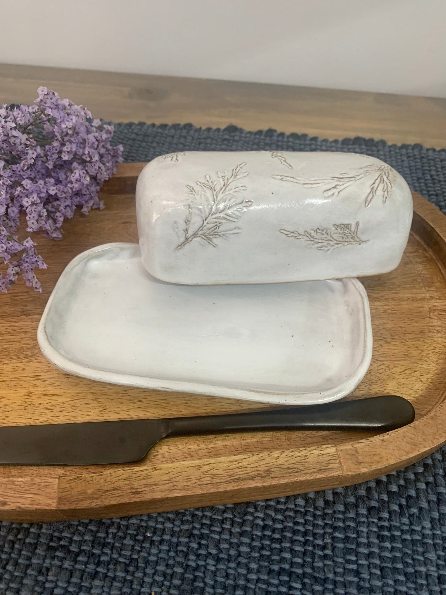 White artisan ceramic butter dish with lid and knife on a wooden tray with purple flowers.