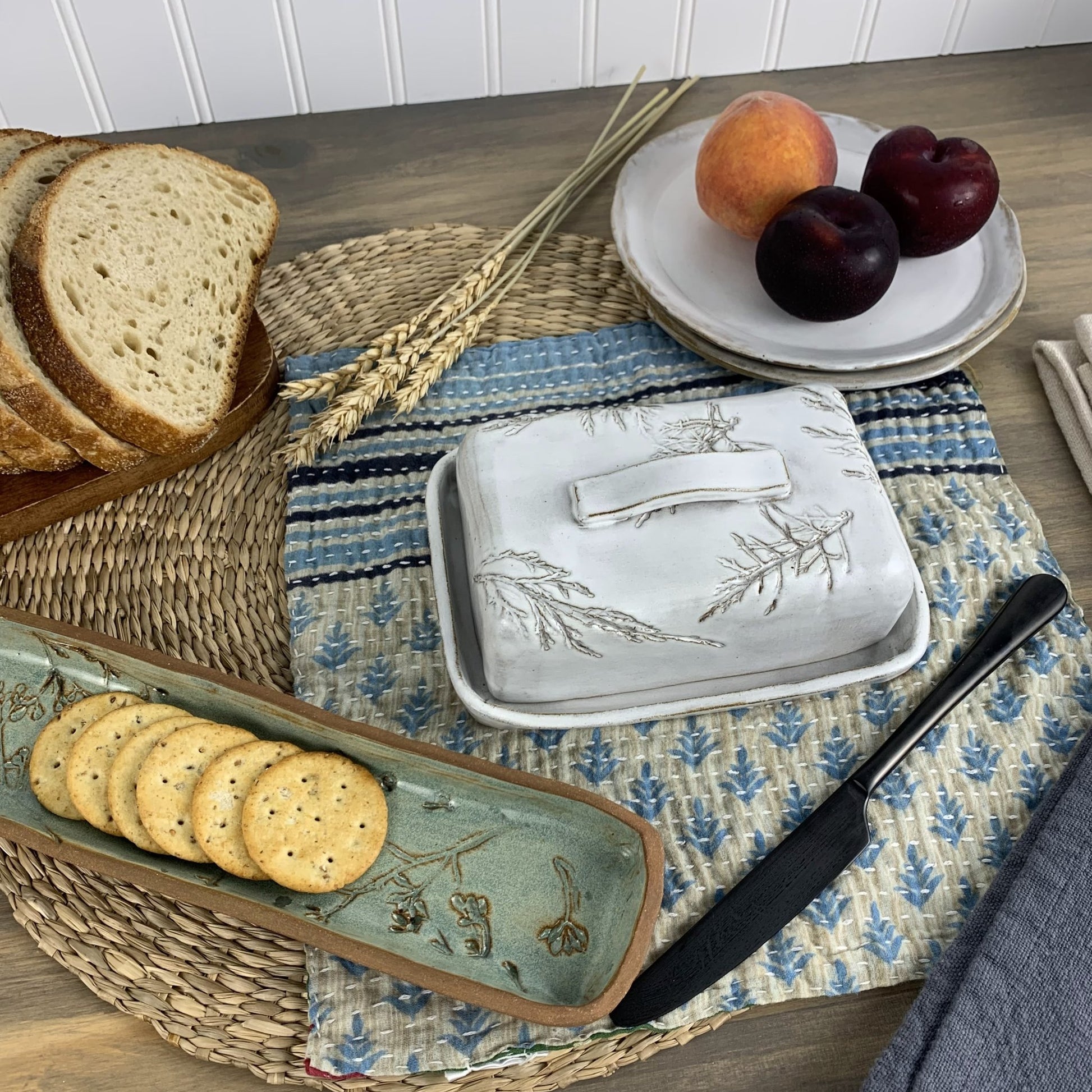 Handmade ceramic butter dish with crackers, bread, and fruit on a table with a blue and white checkered cloth.