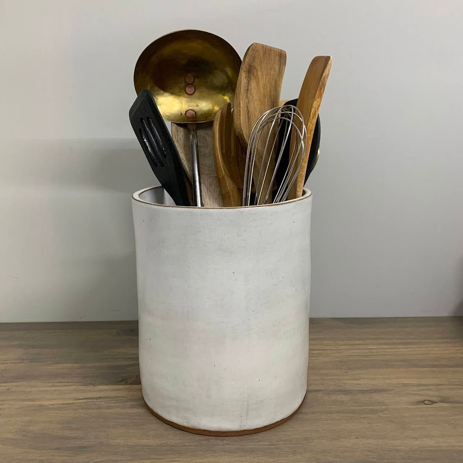 White handmade ceramic utensil holder with kitchen utensils on a wooden surface.