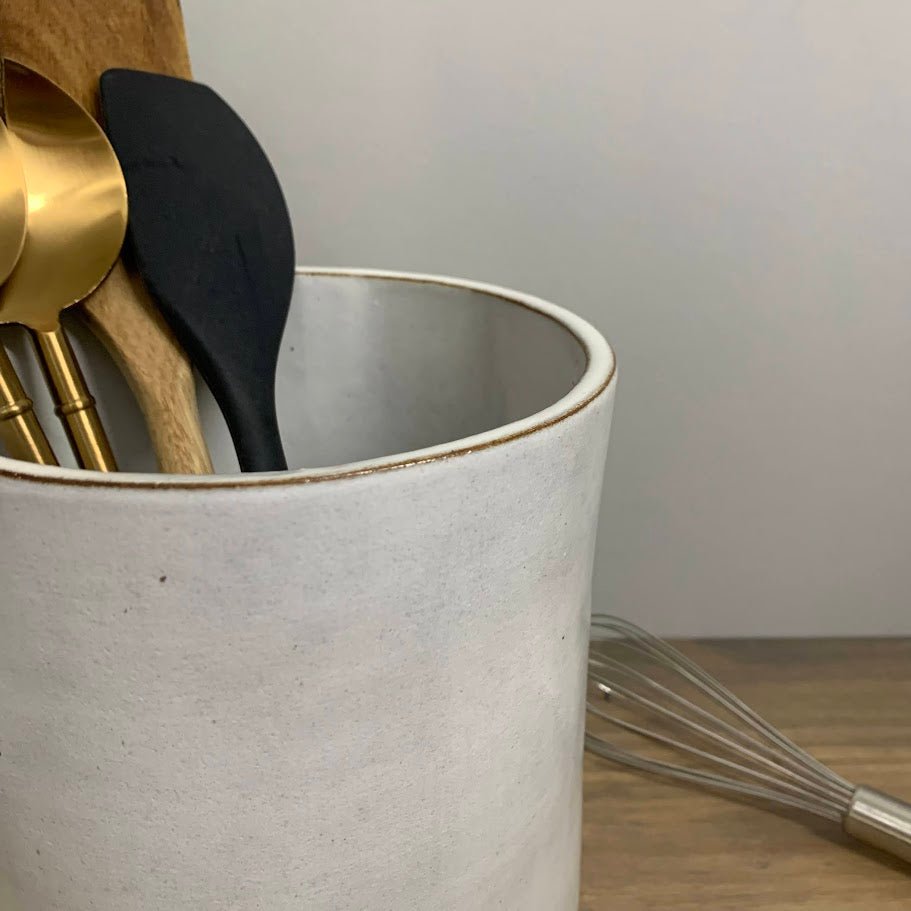 Kitchen utensils including a black spatula and gold ladle in a white handmade ceramic vase on a wooden surface.