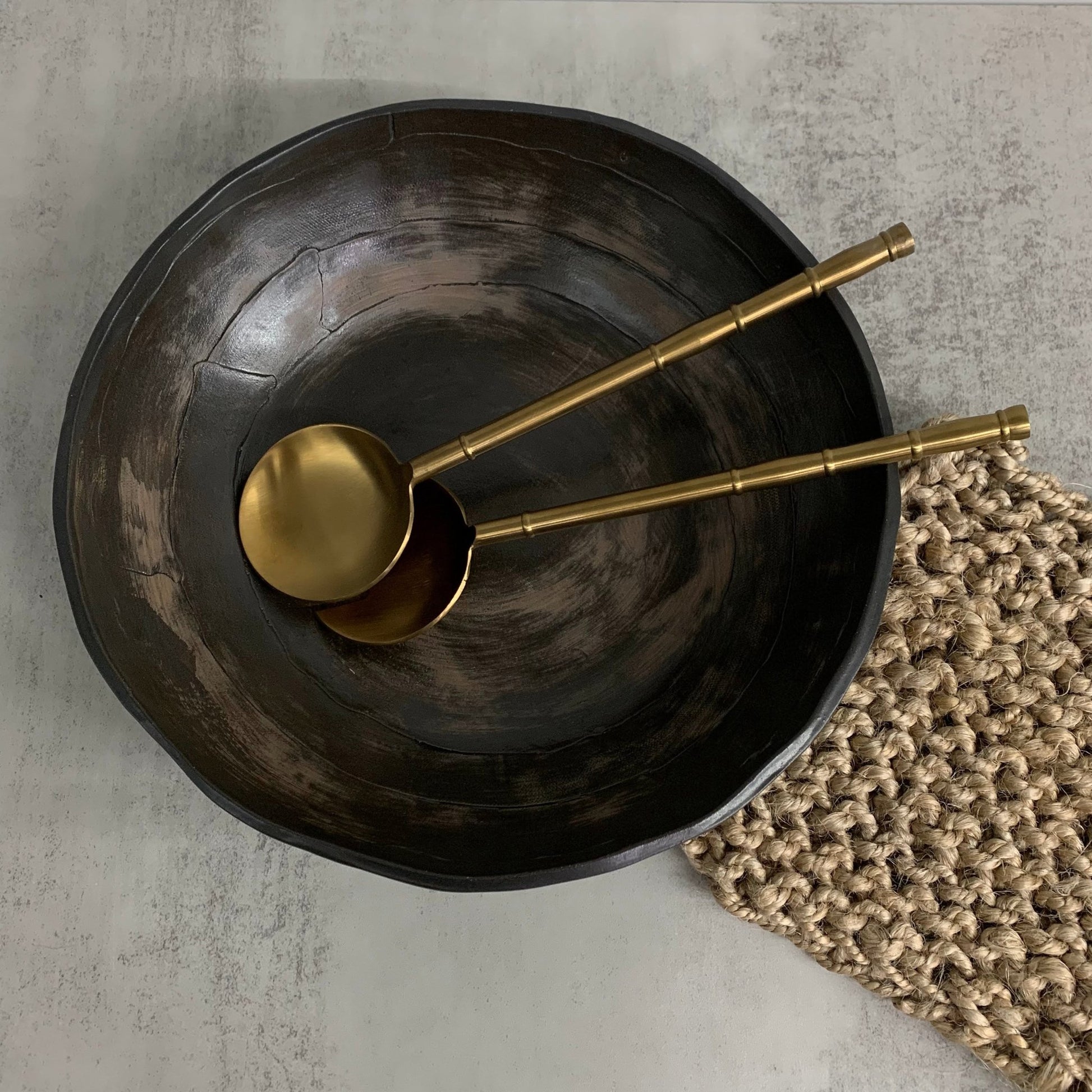 Two gold spoons on a black stoneware bowl with a textured mat on a gray surface