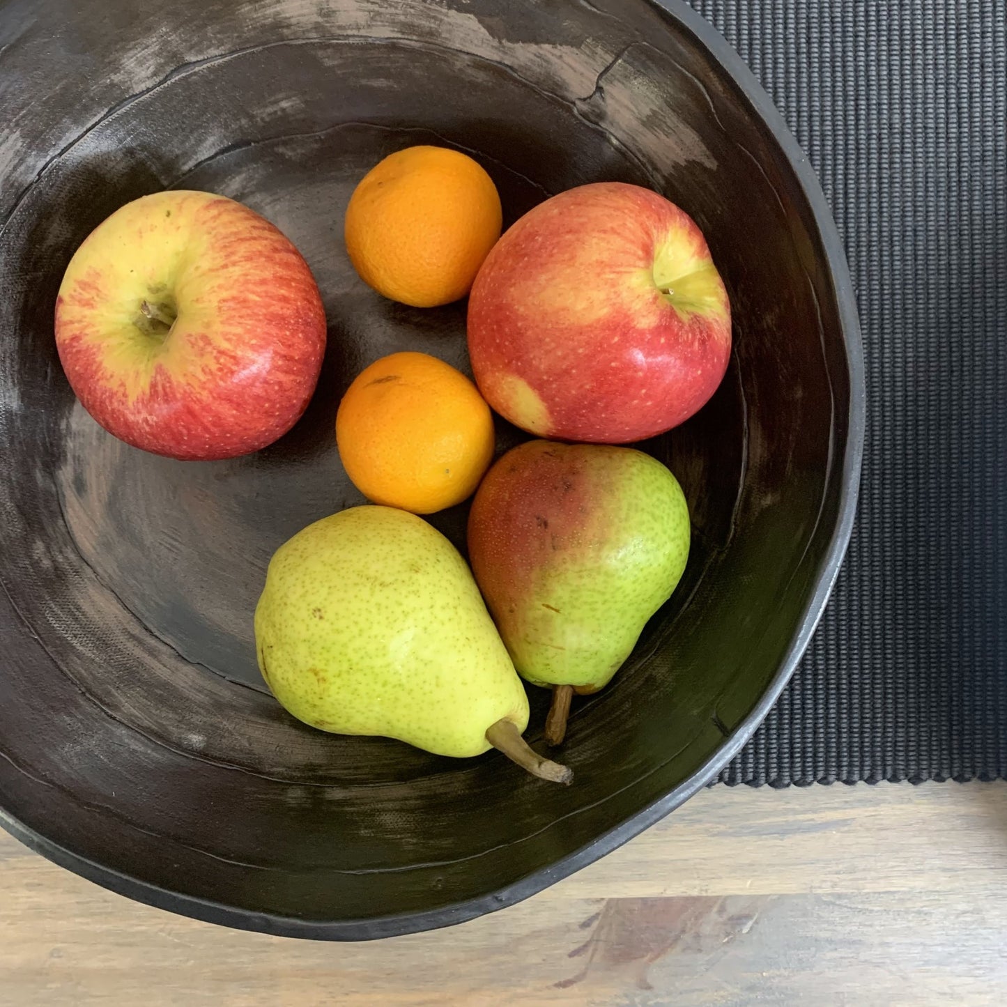 Fruit including apples and pears in a black handmade ceramic bowl on a textured surface.
