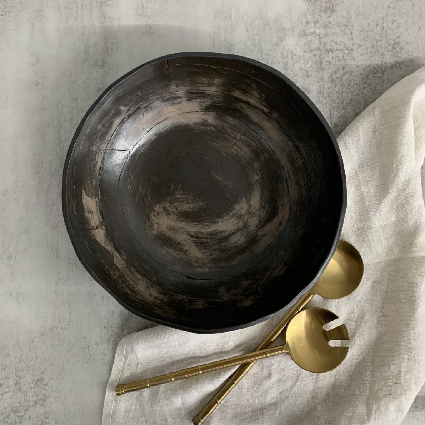 Black handmade ceramic bowl with gold utensils on a light fabric surface
