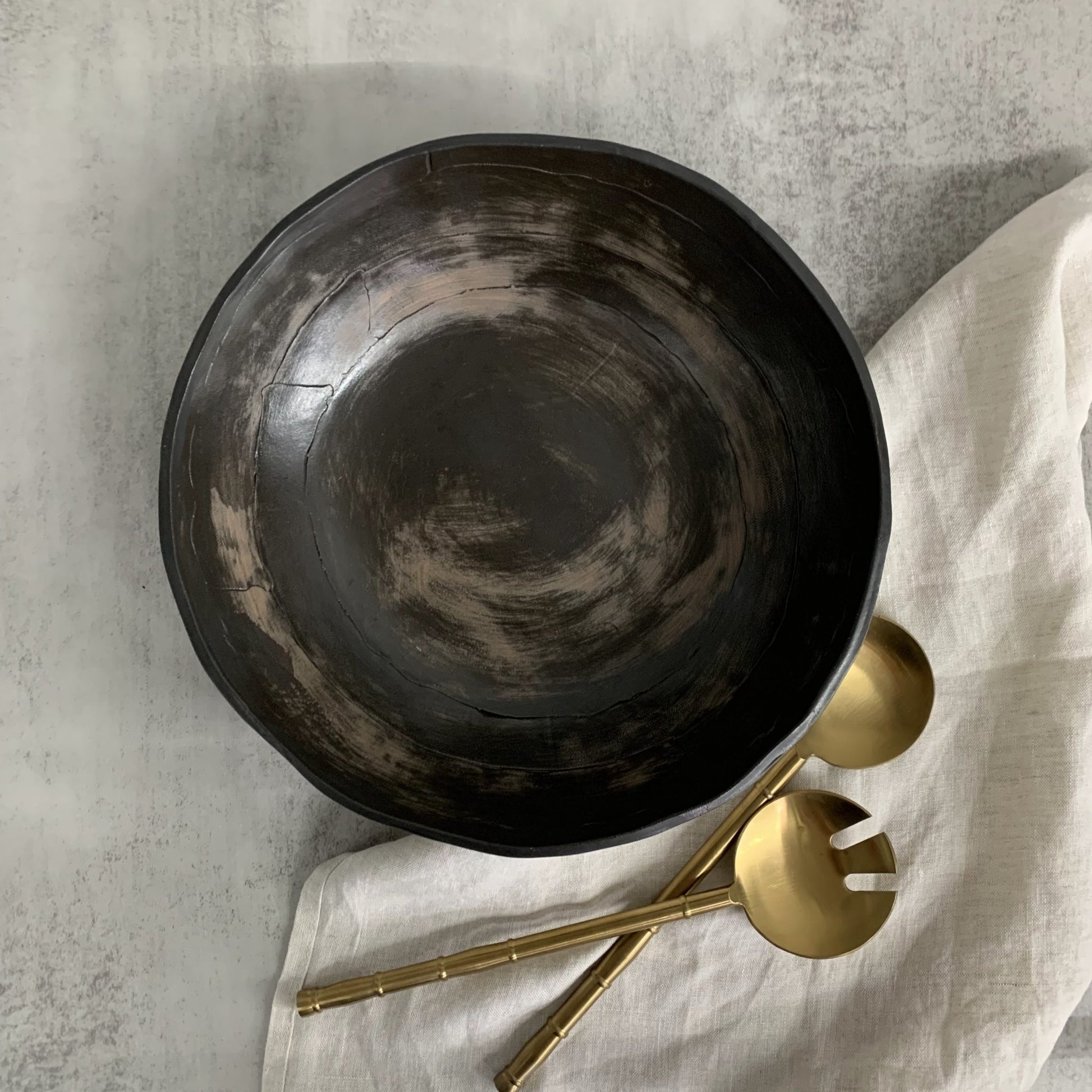 Black handmade ceramic bowl with gold utensils on a light fabric surface