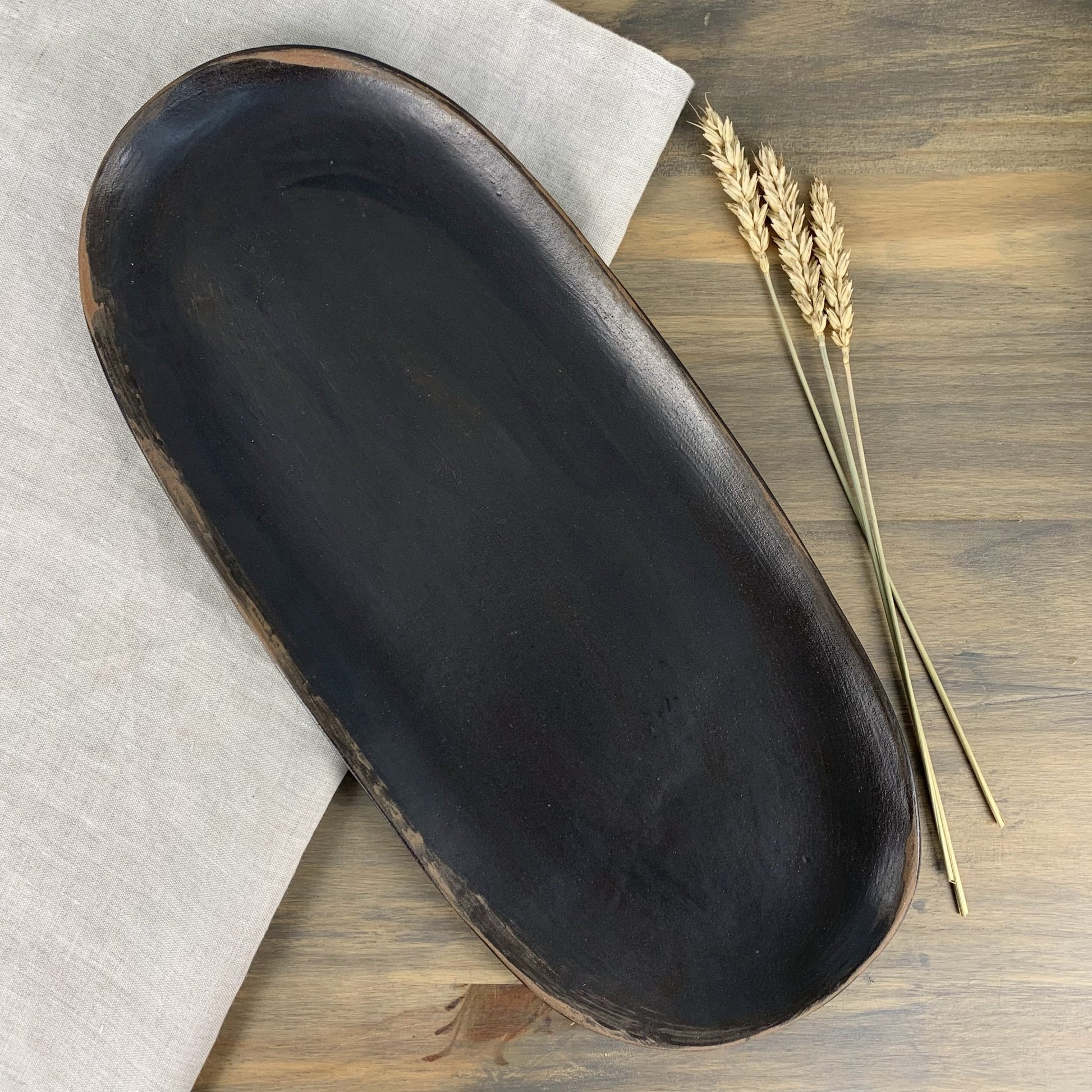 Black handmade ceramic tray on a wooden surface with a piece of paper and dried wheat stalks.