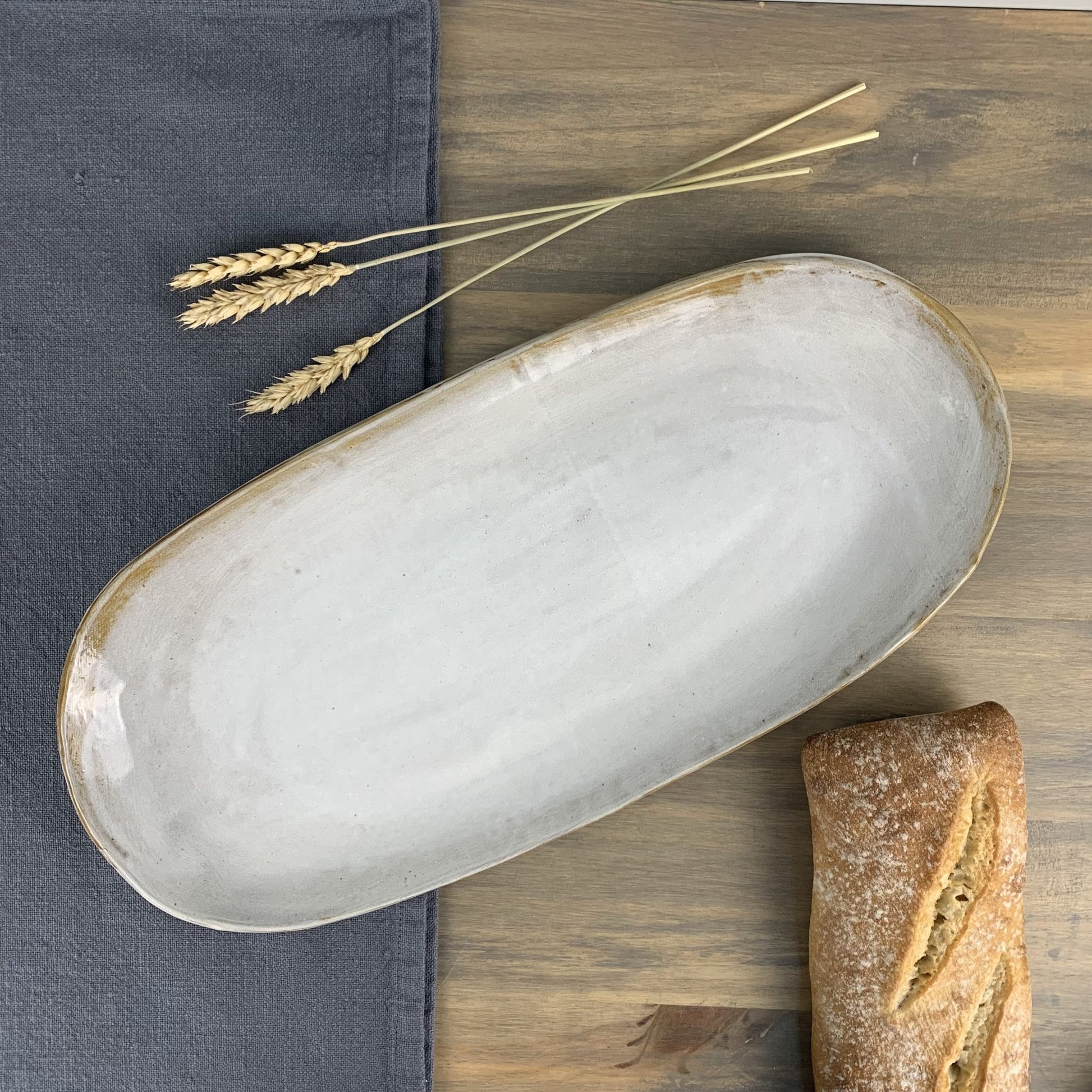 White ceramic stoneware oval shaped platter on a wooden surface with bread and wheat ears.