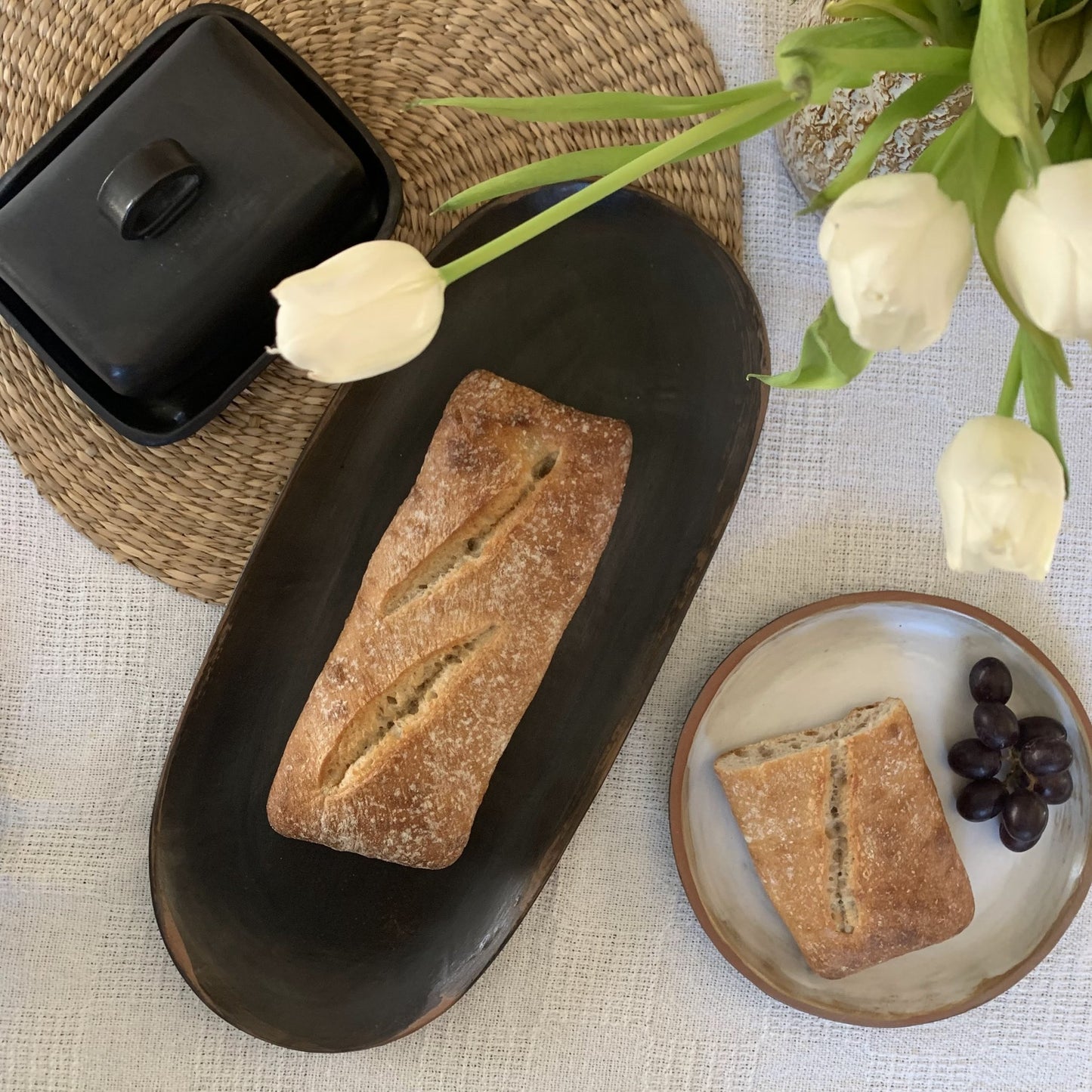 Loaf of bread on a handmade black ceramic plate with white tulips and grapes on a light surface