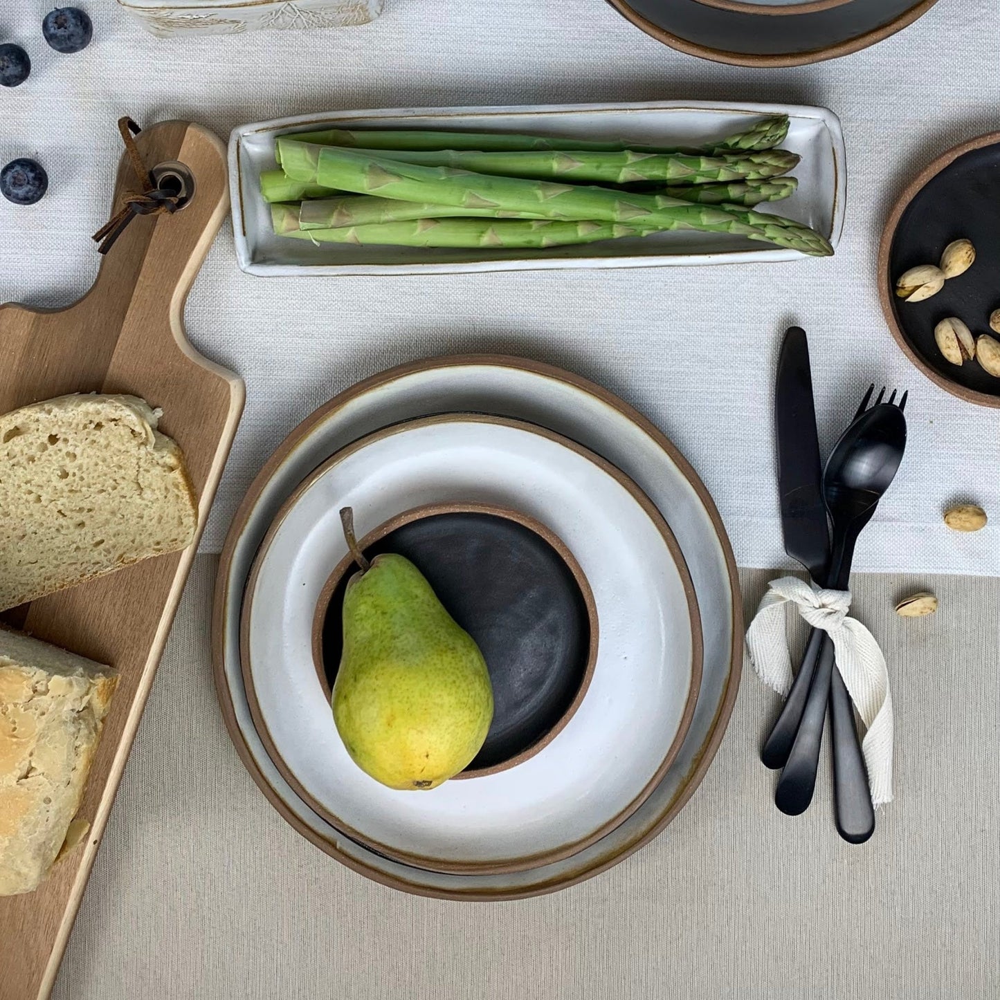 Table setting with handmade ceramic dinnerware, bread, asparagus, and a pear on a light gray surface.