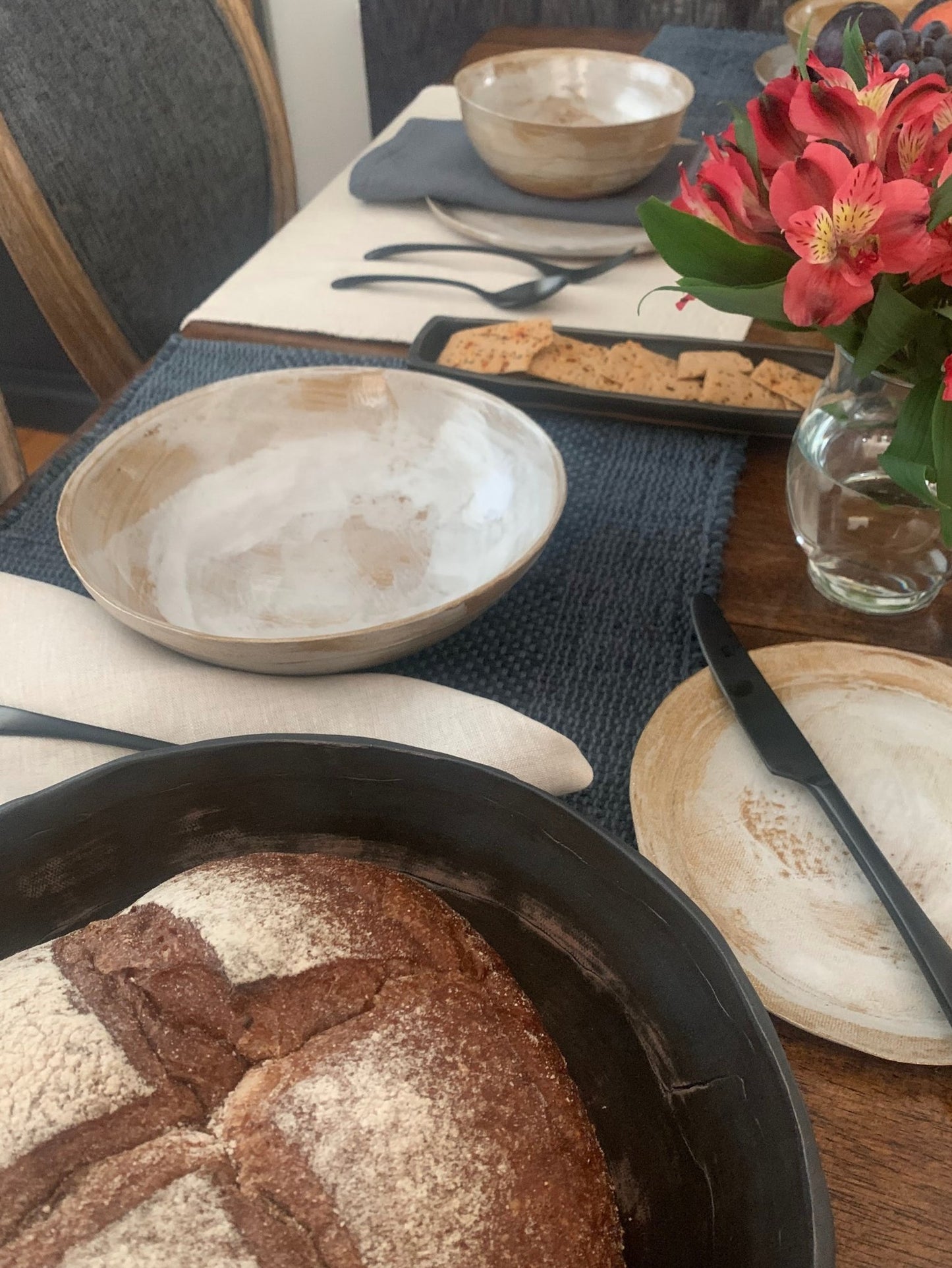 Loaf of bread in a handmade ceramic bowl on a table with flowers, and other items.
